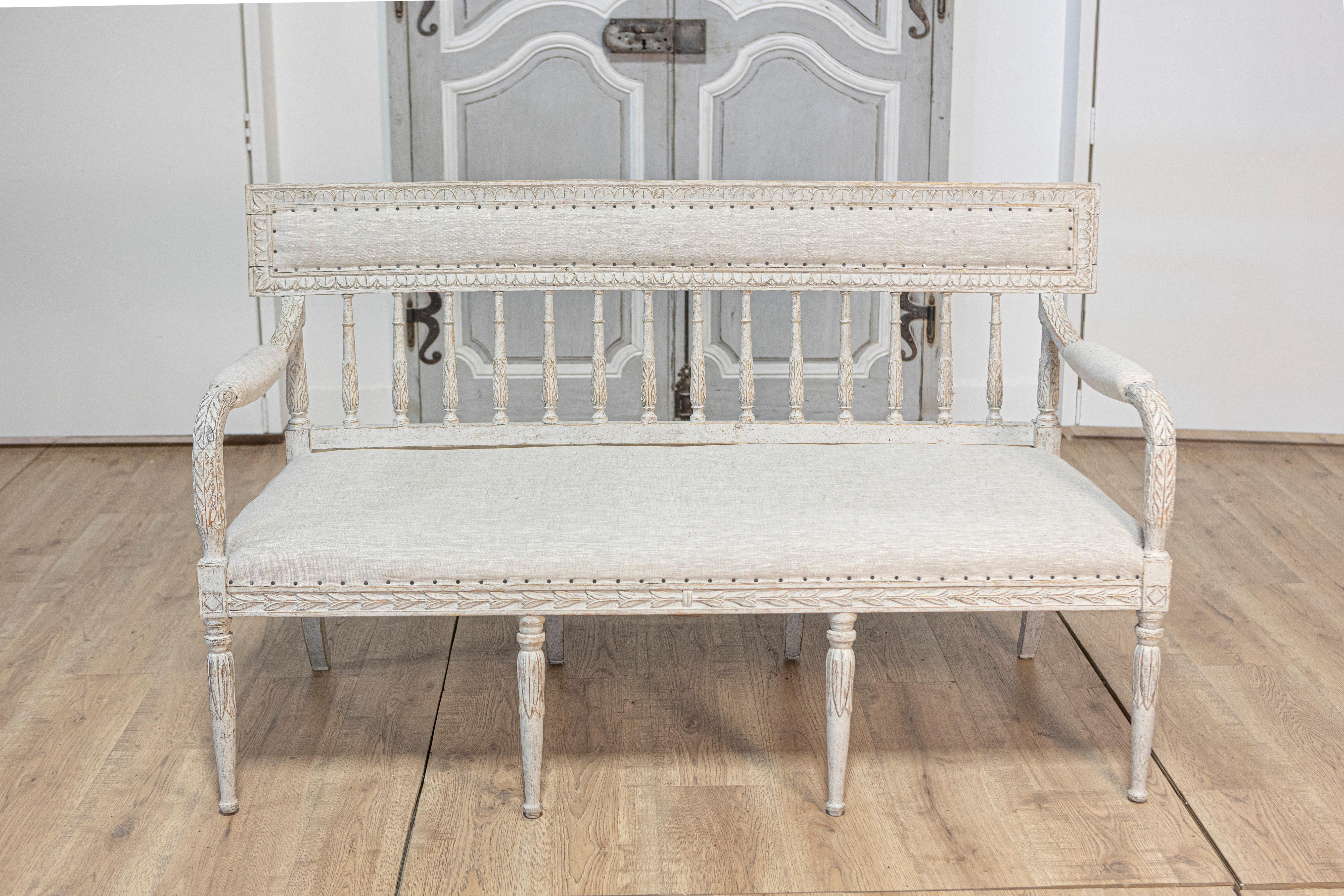 This 20th-century Swedish Gustavian-style settee exemplifies the elegance and simplicity of Scandinavian design. Crafted from pine, the settee features a soft, distressed white finish that enhances the intricate carvings along the arms, backrest,