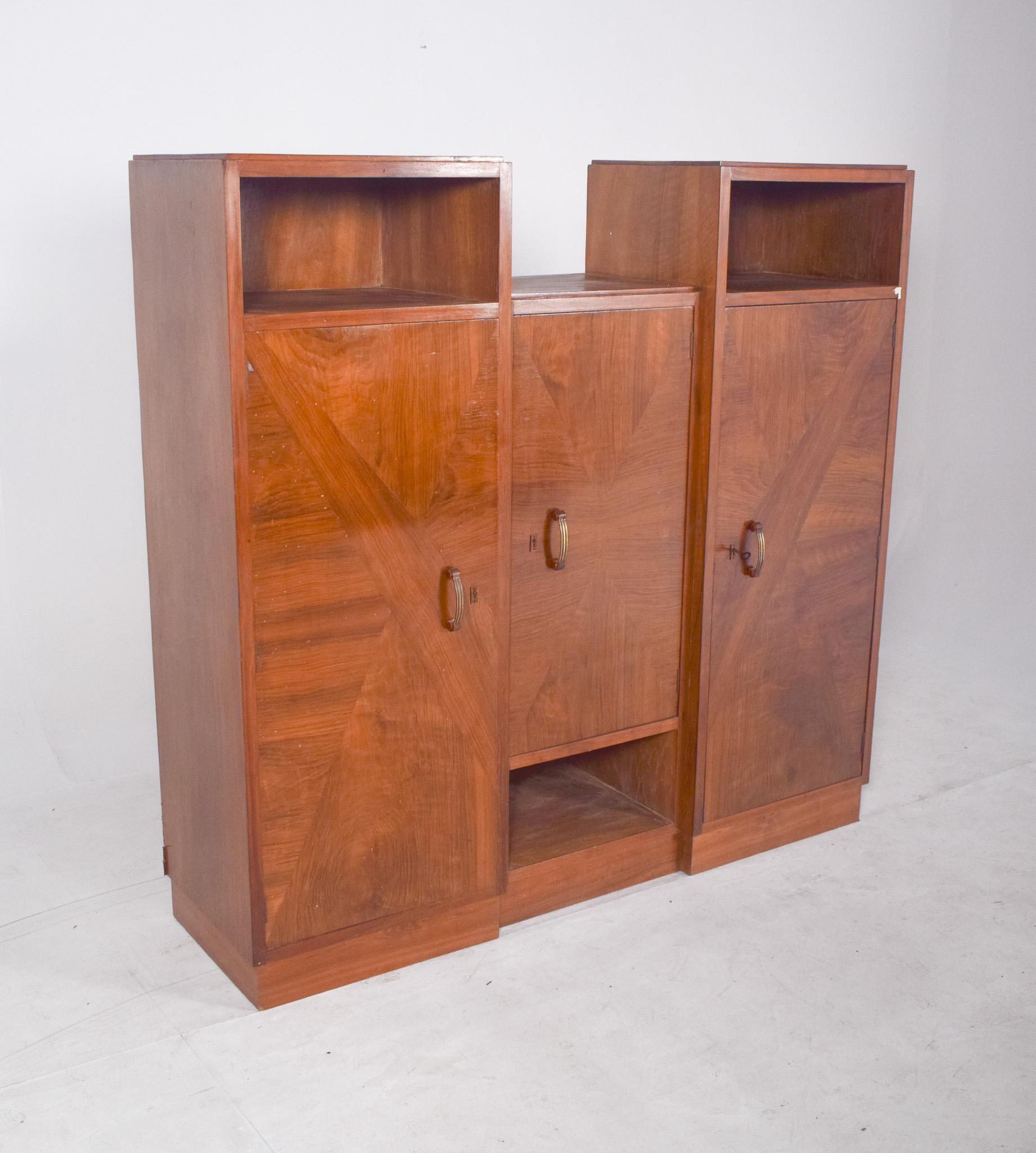 Bibliothèque à trois portes. De conception symétrique et robuste, il comporte trois compartiments principaux avec portes, ainsi que des niches ouvertes en haut des côtés. Les portes sont dotées de poignées verticales et de détails de marqueterie aux