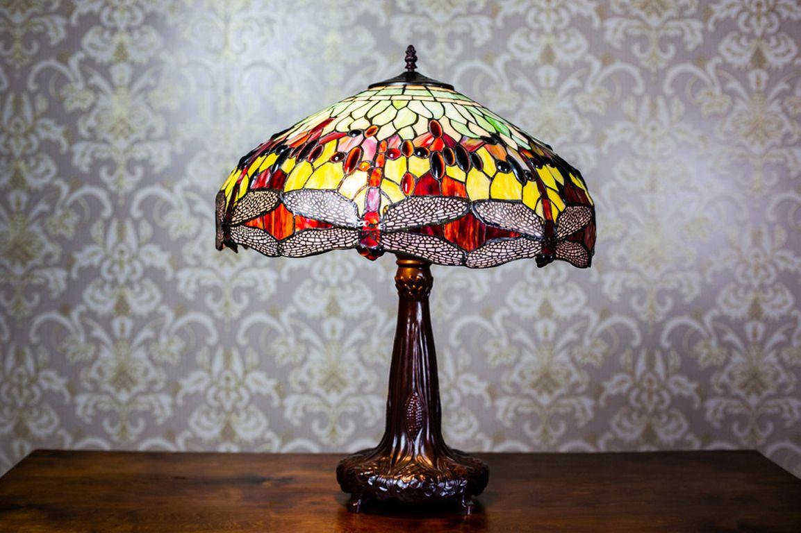 We present you a lamp with a base of painted aluminum, which imitates bronze, and a stained glass shade.
The shade is made of multi-color glass with a motif of dragonflies.
Three light bulbs with a big base are the light source.

Presented item