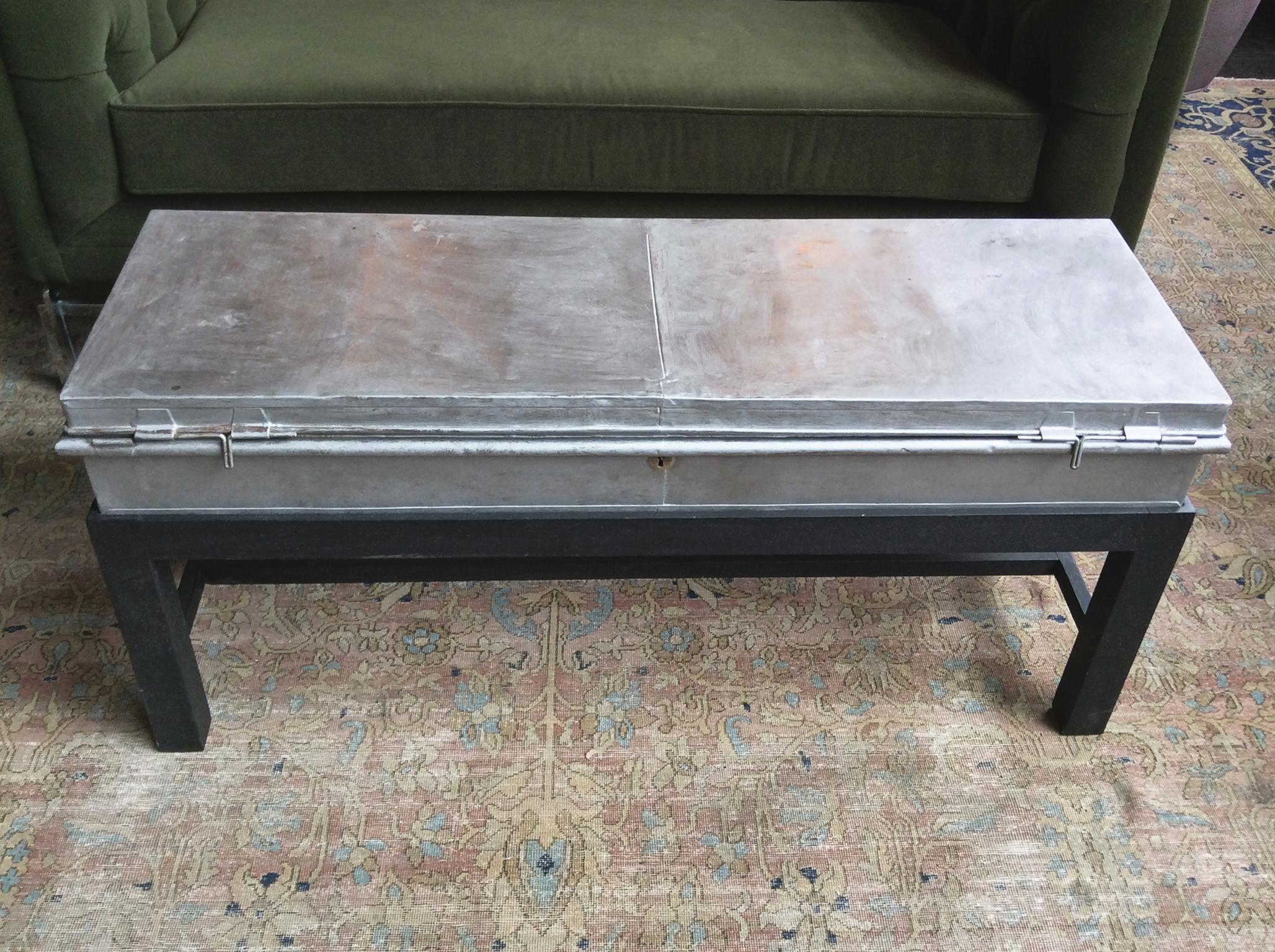A versatile industrial coffee table comprised of a tin box top and a wooden base. Hardware includes a keyhole, two barrel bolts, and two handles at either end. A fog-white wash covers the metal, giving it a purposely weathered look. The box's hinged