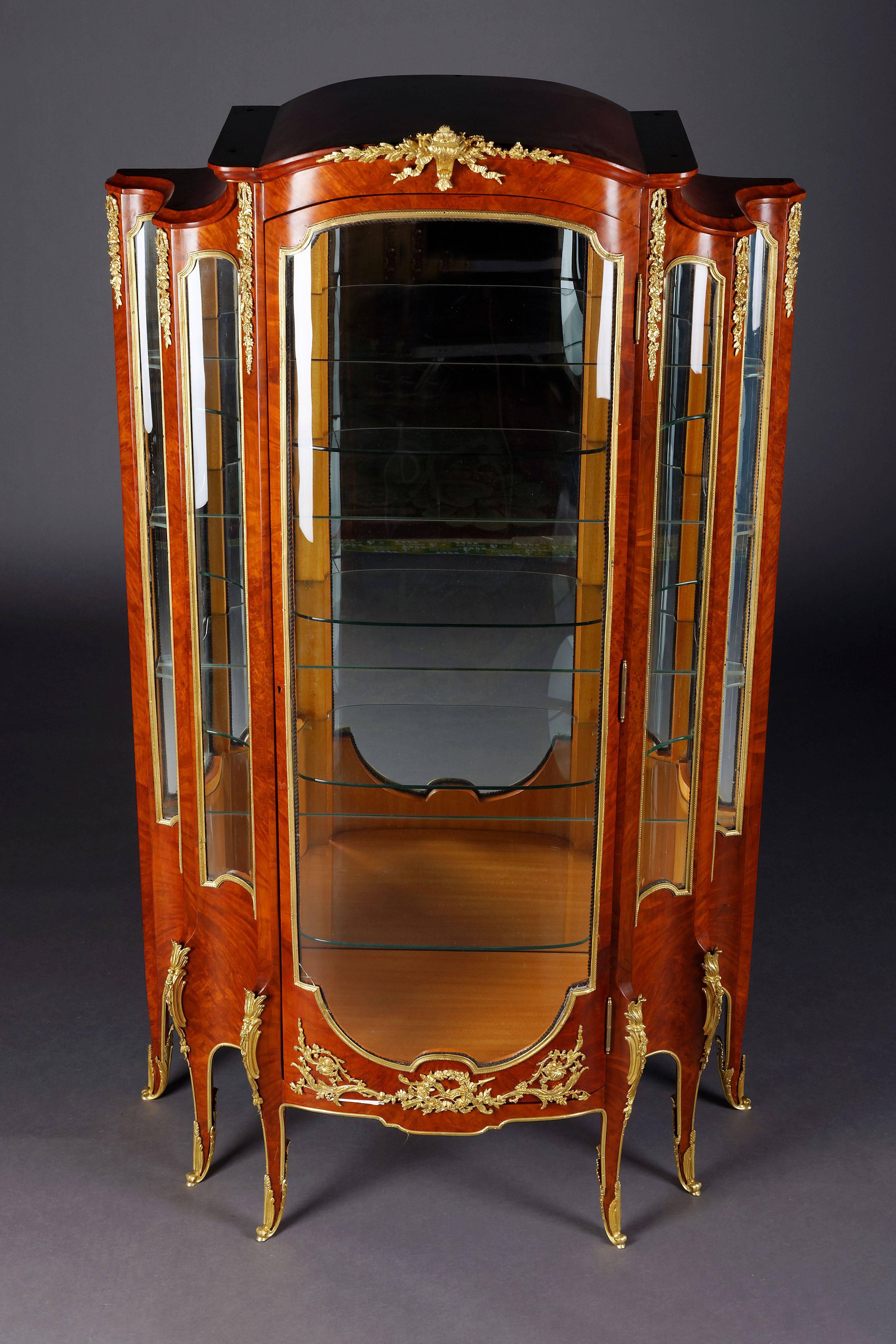 Majestic French vitrine in transition style.
Root veneer on solid conifers. High-angled, one-door, cambered and three-sided glazed body on tall, slanting, curly feet in sabots. Three-sided scalloped, cambered, frame. Rich in extremely finely