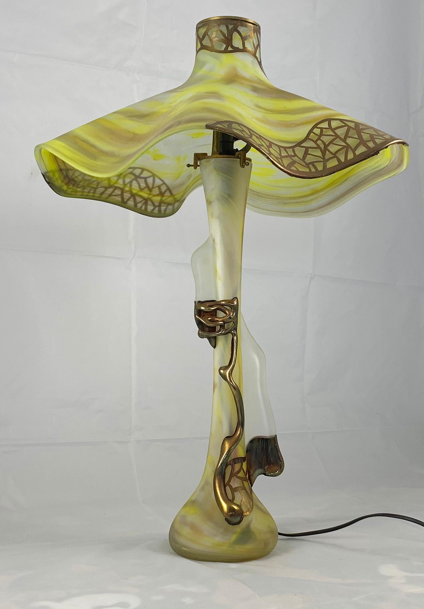 An unusual art glass table lamp in Art Nouveau style. In the manner of Daum. The slender baluster body with long tapering neck supporting a large shade. In green glass with striation decoration picked out with applied base metal.