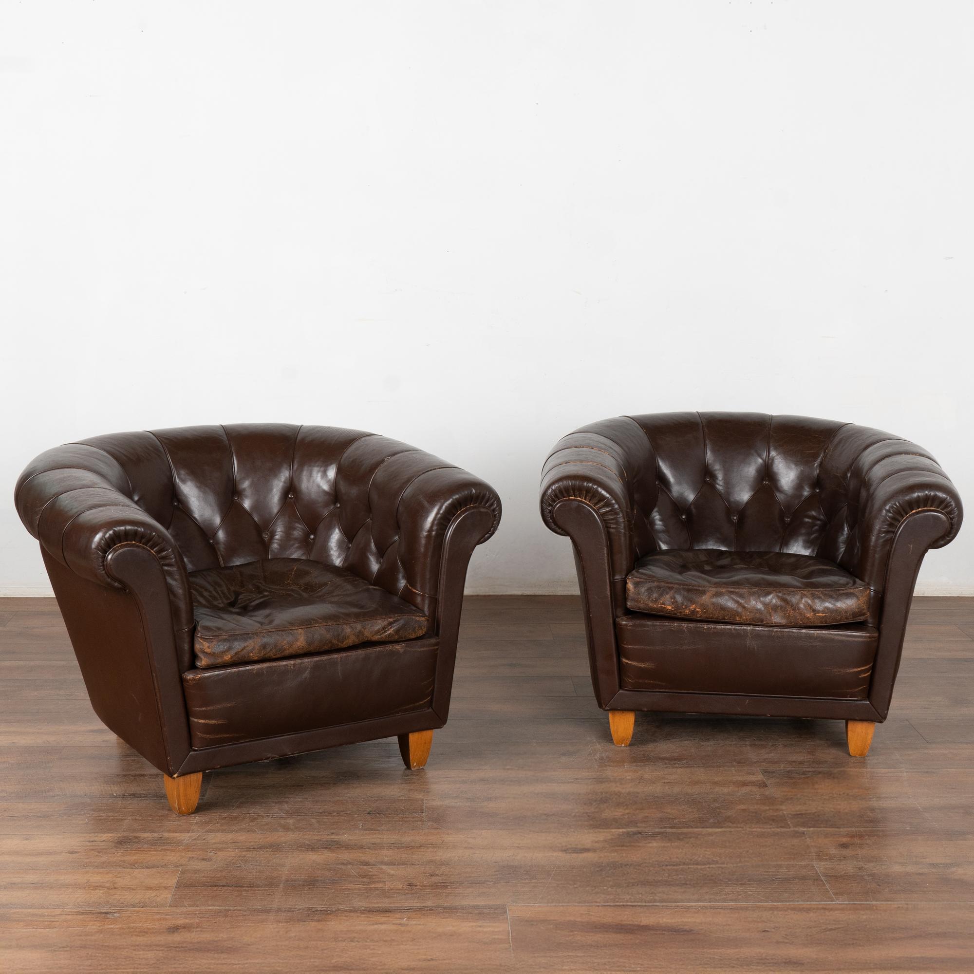 This pair of vintage leather club chairs are welcoming with barrel shaped arms and tufted backs with self covered buttons and piping.
This pair shows signs of typical age and wear to the dark brown leather including scratches, stains, scuffs,