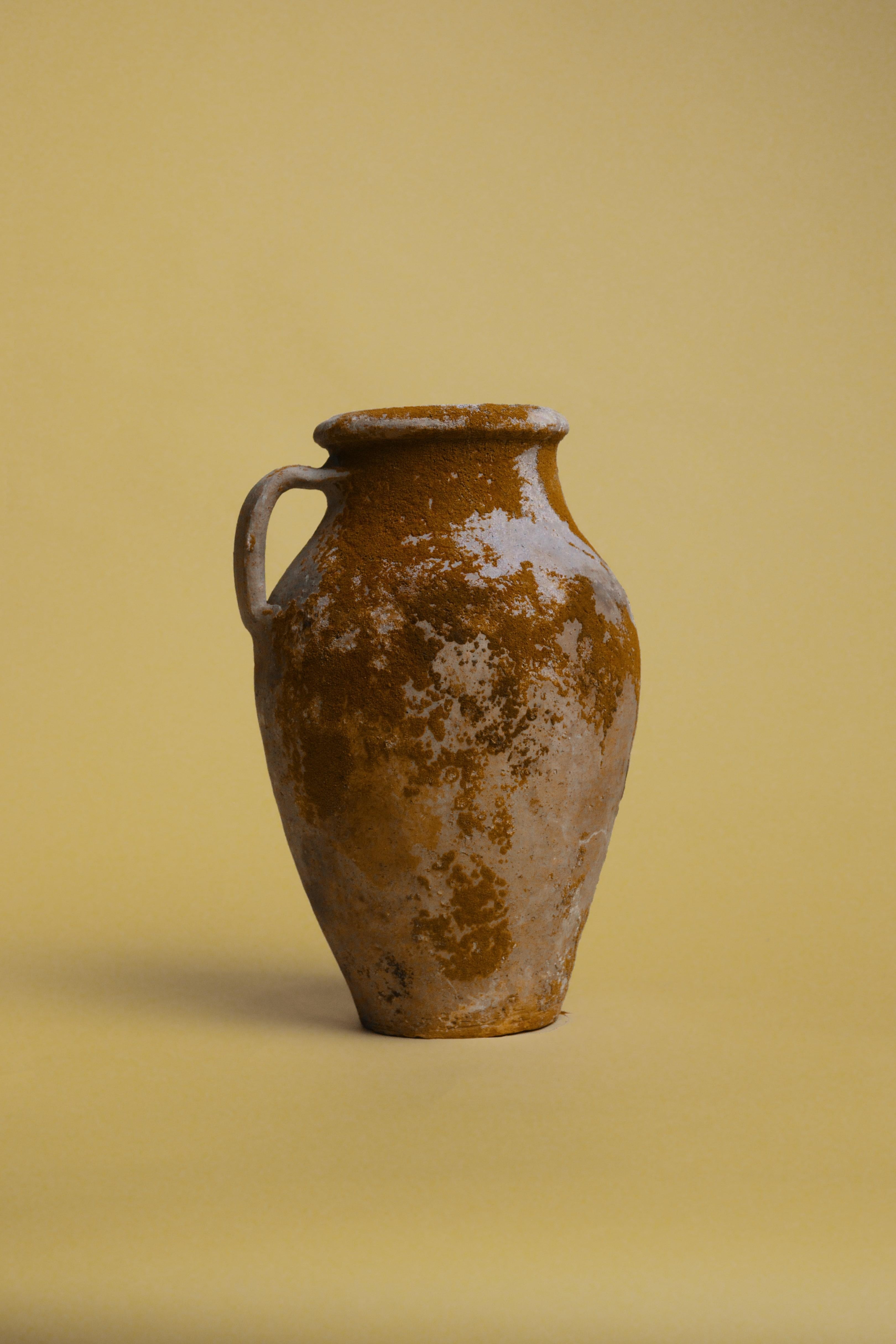 Introduce calidez y textura en tu espacio con este jarrón vintage de terracota con reflejos ocres, un sorprendente ejemplo de la artesanía de la cerámica mediterránea. Este recipiente de barro rústico, fabricado a mano con técnicas tradicionales,