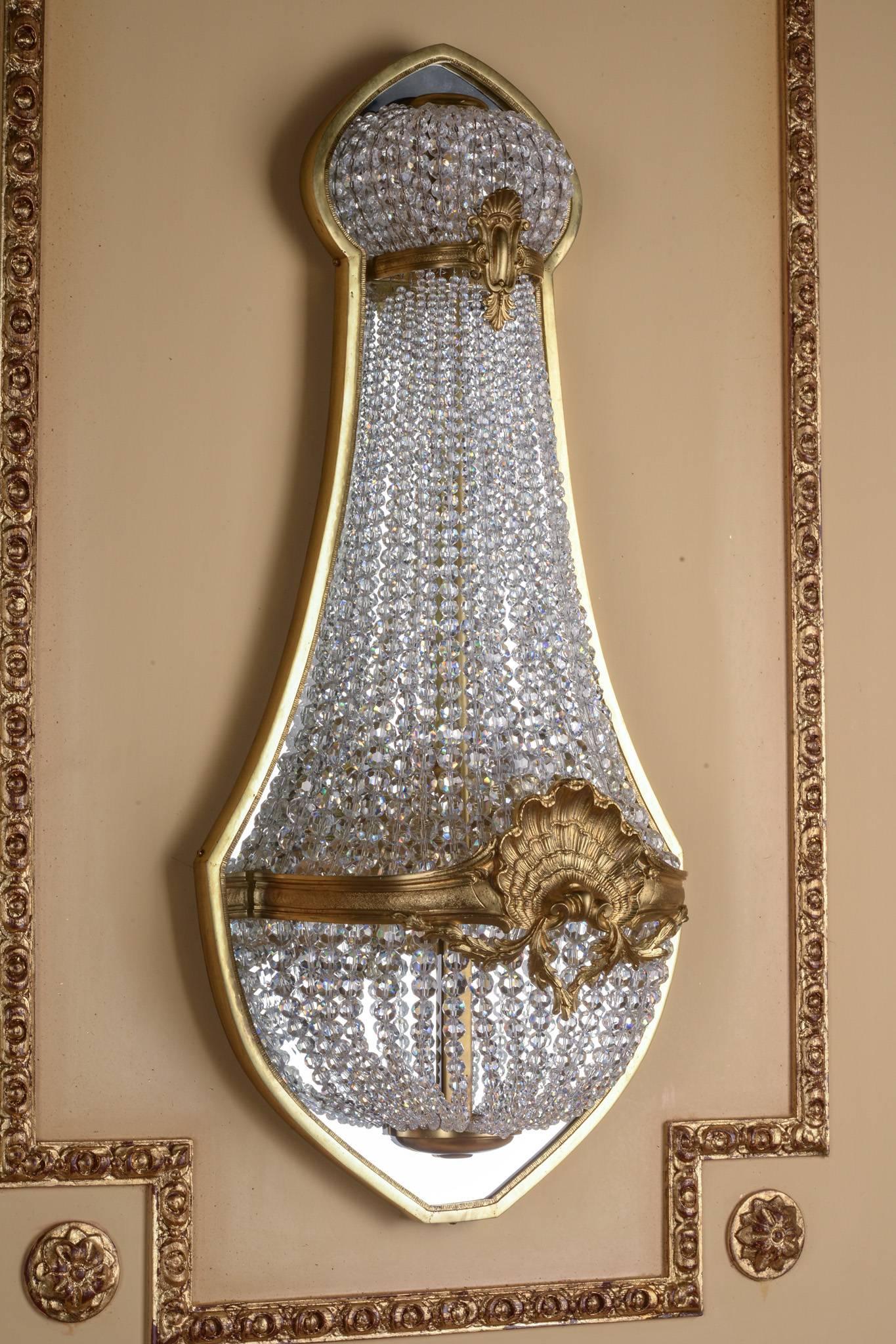 A classicist wall sconce with finely chiselled bronze. Basket-shaped body made of handcut French ball prisms in the course of broad, ornamental-reliefed ring connected centrally placed shell. The finely ground crystals break and reflect the light