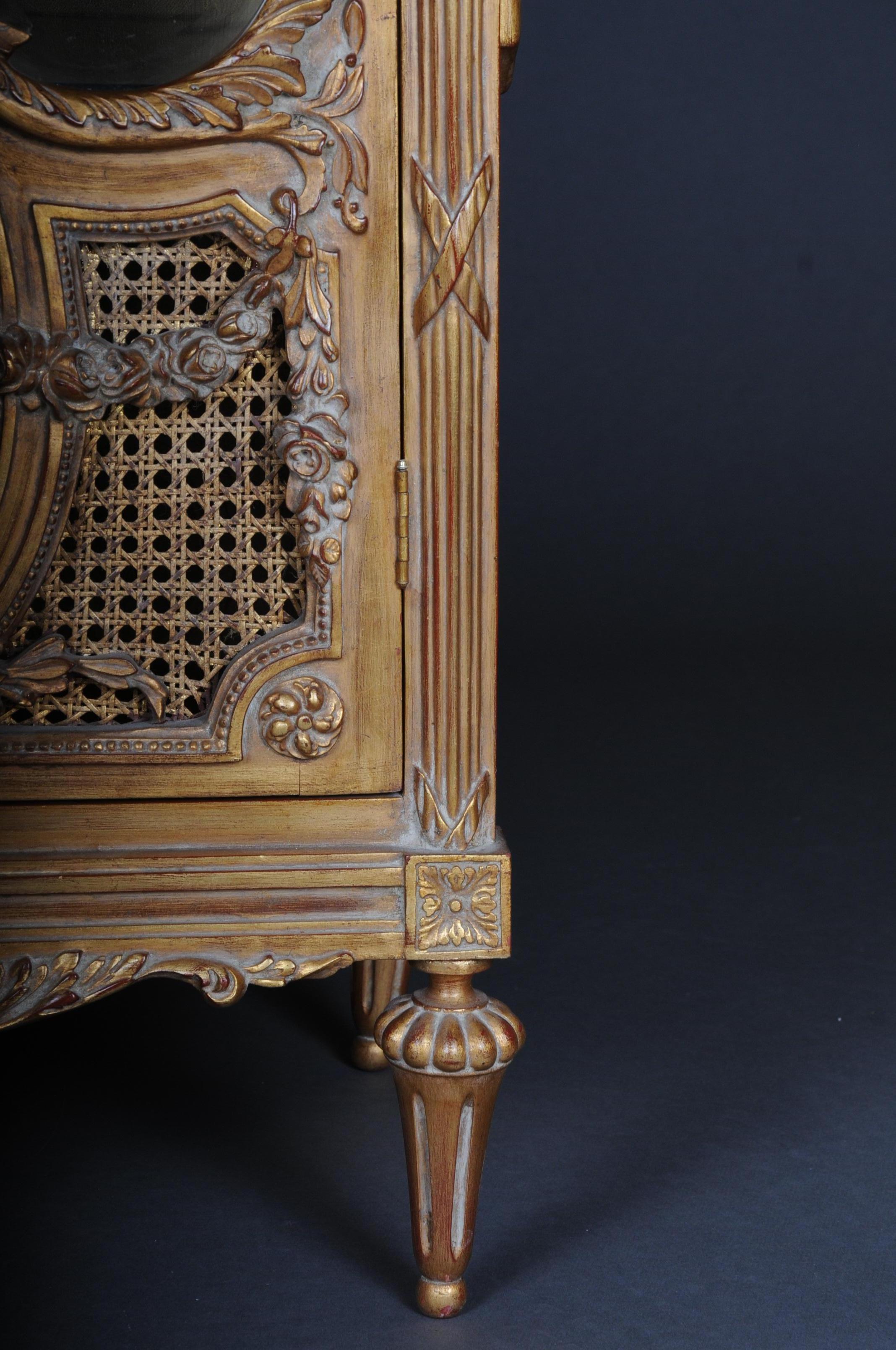 Solid beech, gilded. Rectangular, three-sided, faceted beveled, one-door body on conical legs. The door is partially provided with wickerwork. The front, corners and sides are decorated with carved, classic elements such as garlands and bows.