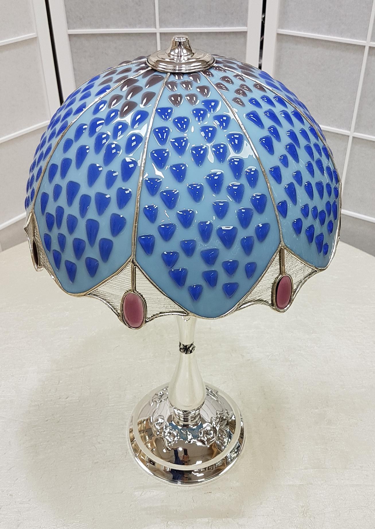 Sterling silver lamp with a smooth shaped steam with a small decoration in the middle. The base is embossed and chiselled completely by hand with floral designs. 
The lamp cap is in polychrome glass made in Murano in light blue, gray, blue and