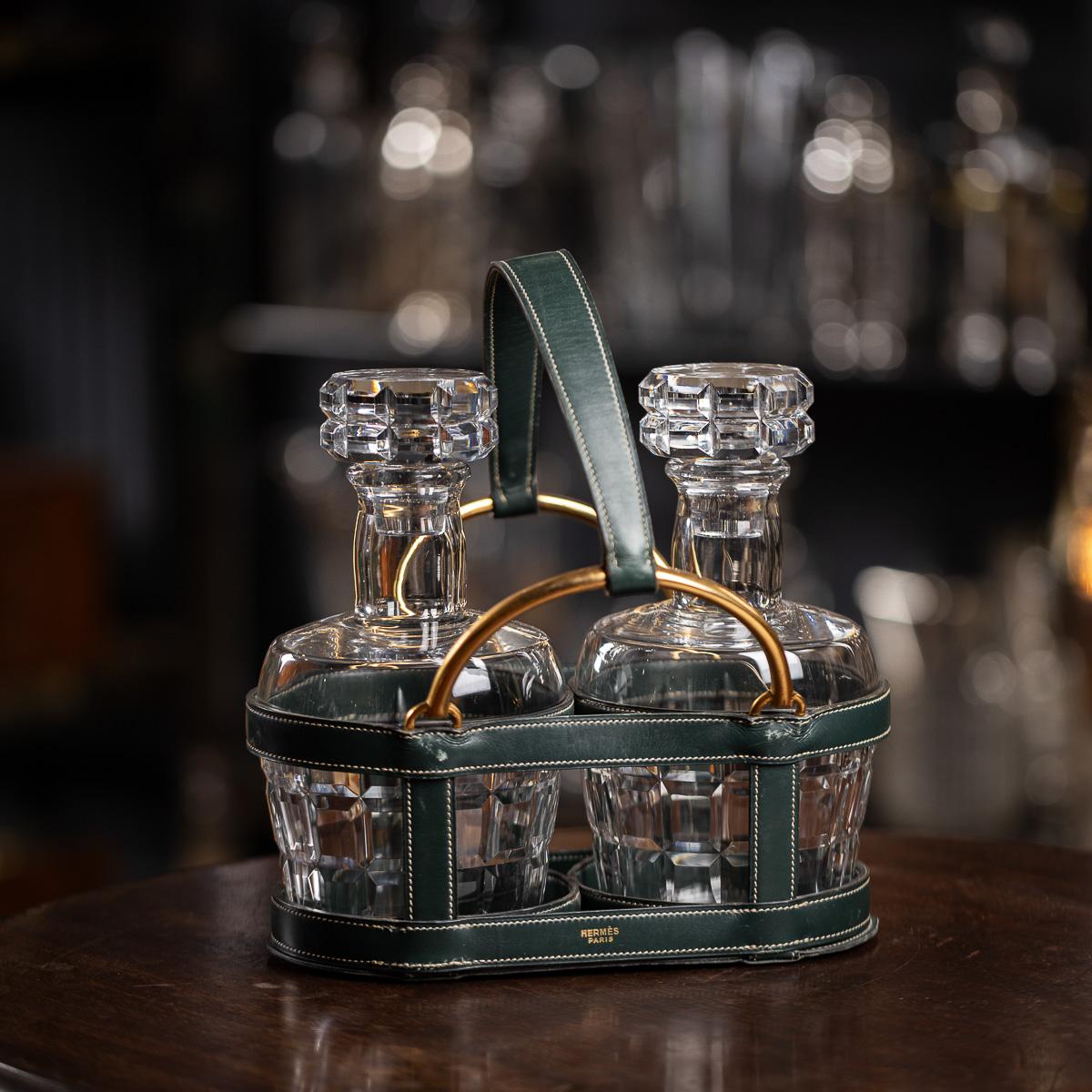 Un double porte-bouteille français vintage du 20ème siècle en cuir vert avec des détails en laiton, conçu par Jacques Adnet pour Hermès. Il abrite une paire de carafes en cristal de Baccarat et porte le nom d'Hermès estampillé en or sur le cuir. Il