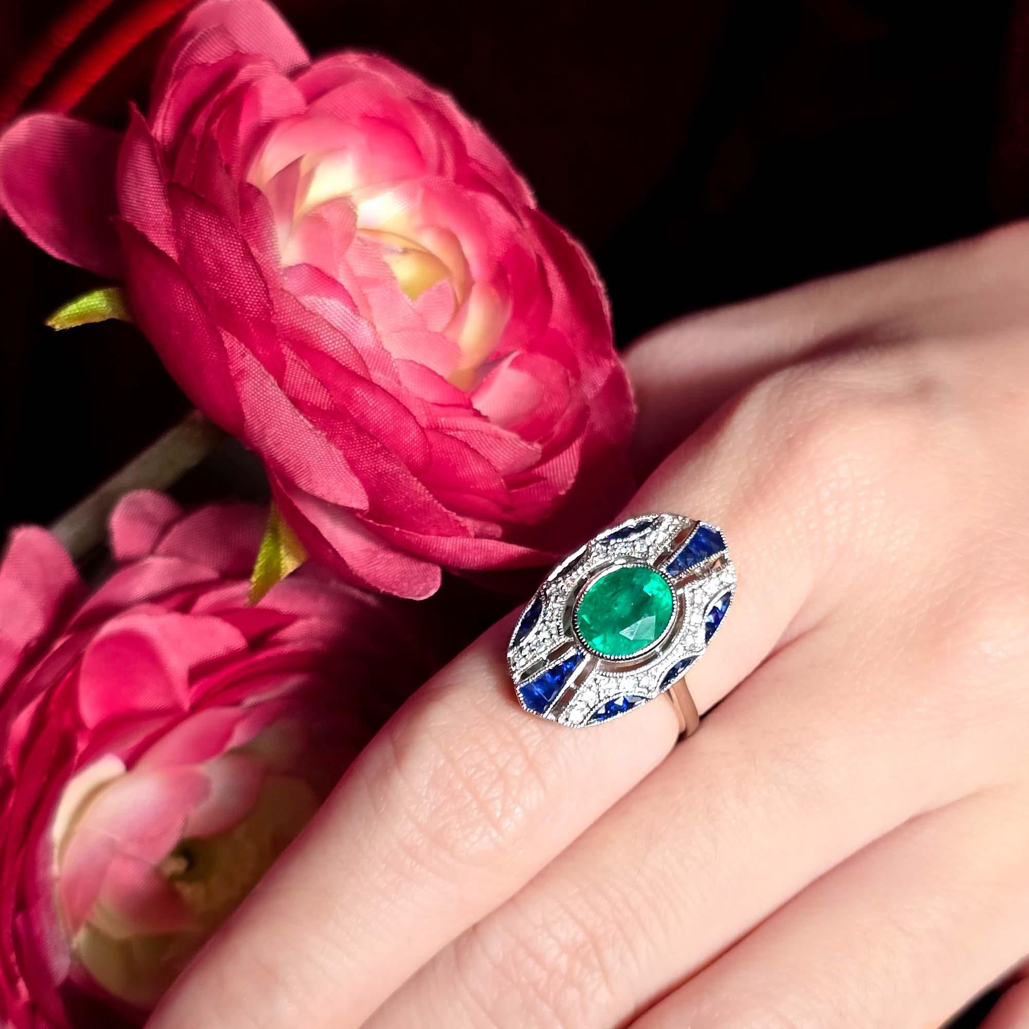 La bague de style Art déco en émeraude, saphir et diamant est ornée d'une magnifique émeraude de 2,1 carats au centre, entourée de diamants étincelants qui rehaussent sa teinte verte vibrante. La monture de la bague est rehaussée de saphirs,