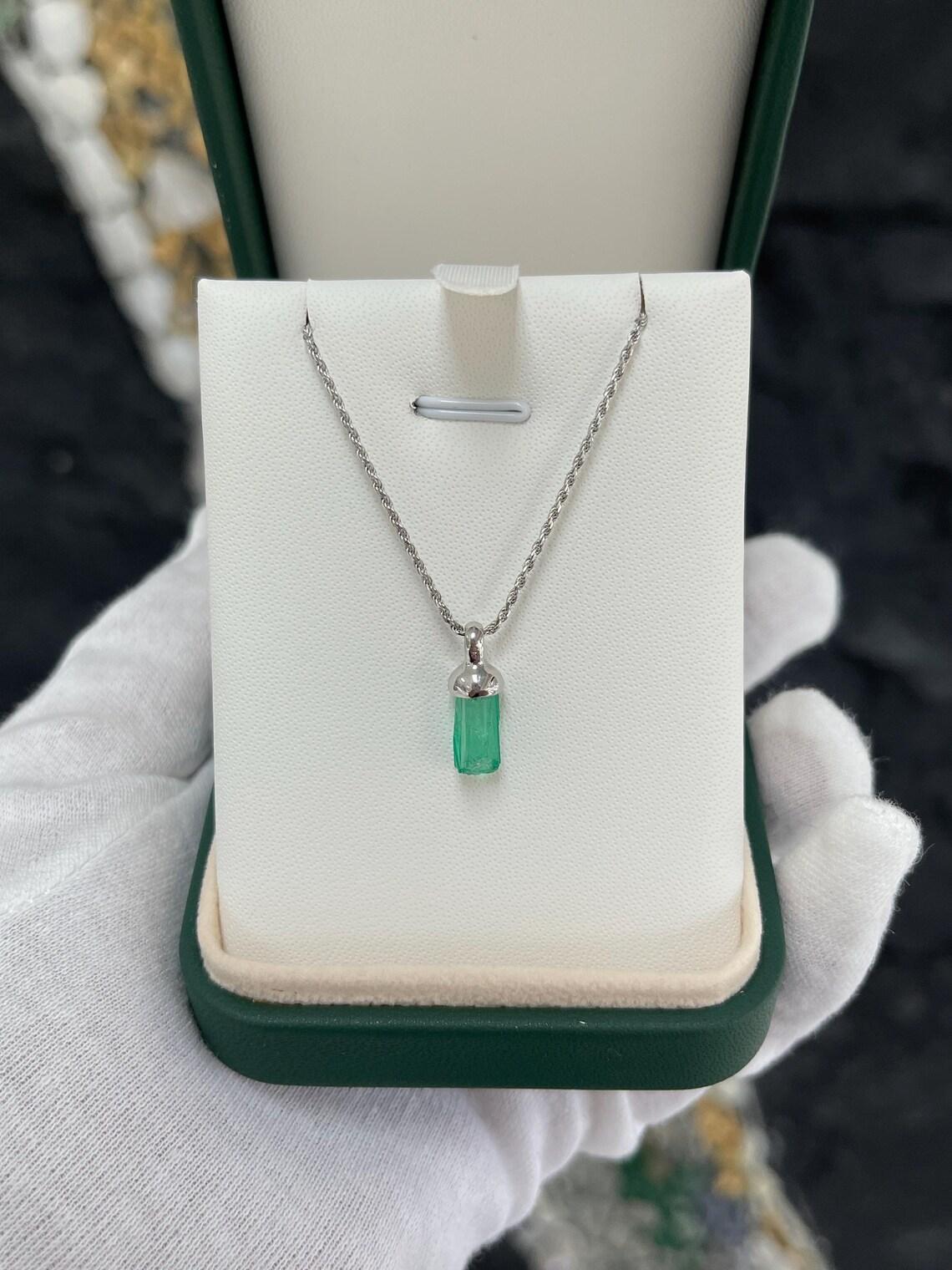 Eleva il tuo stile con il nostro squisito ciondolo a forma di smeraldo grezzo, caratterizzato da uno splendido cristallo di smeraldo terminato da 2,10 carati in un'ipnotica tonalità verde scuro medio, proveniente dalla Colombia. Eleganza senza tempo