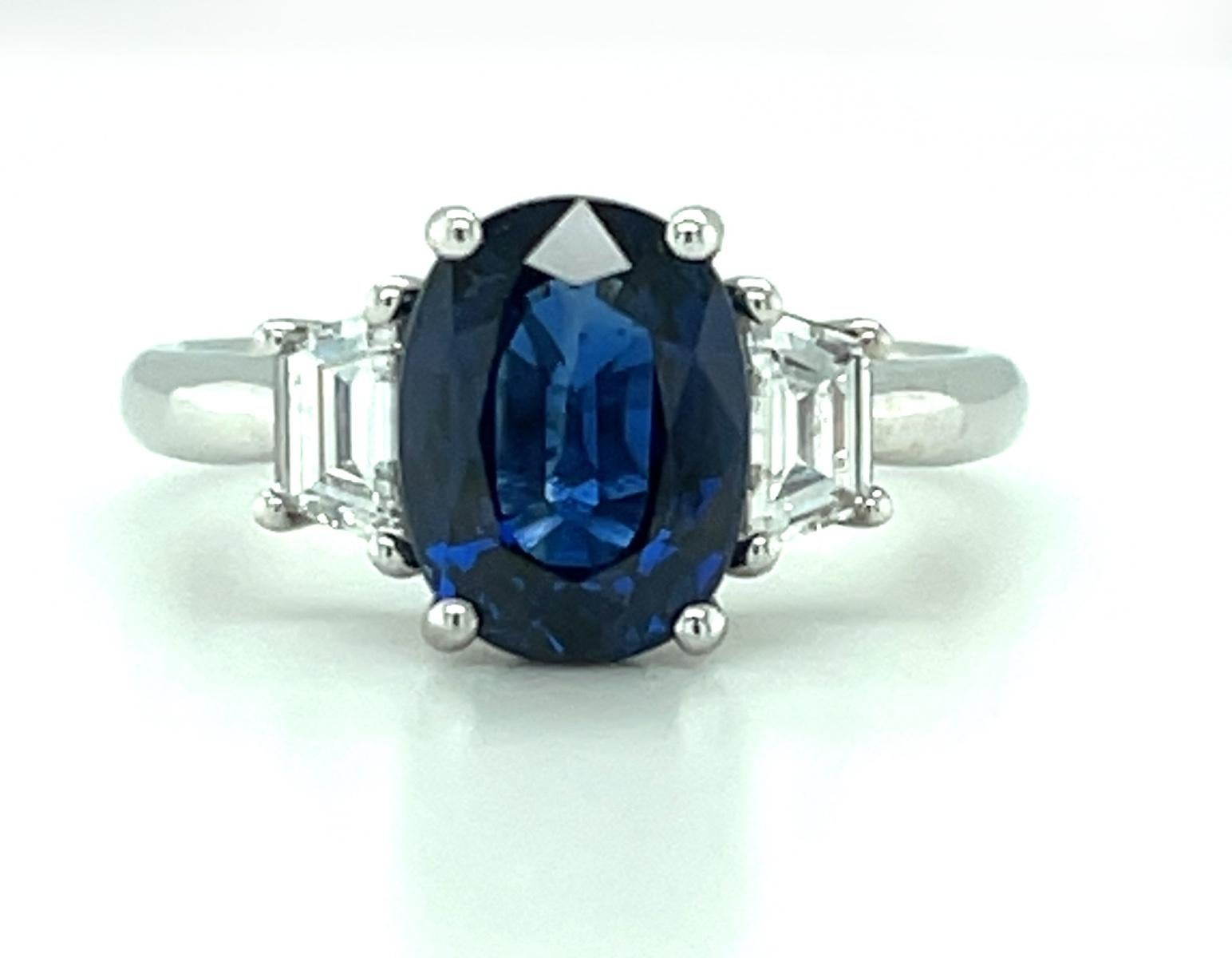 This classic three-stone engagement ring features a beautiful 2.18 carat blue sapphire set in platinum with an exquisite pair of fine diamonds. The sapphire has rich, blue color whose shape is accented beautifully by the trapezoid-shaped side