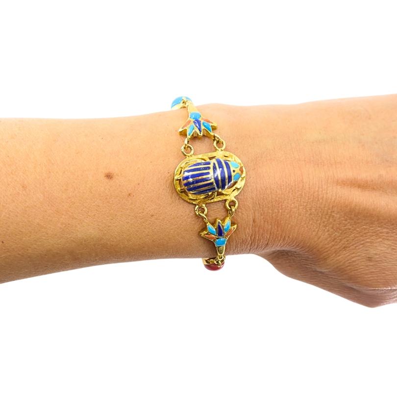 Ce bracelet présente un design néo-égyptien classique, serti en or jaune 21 carats, comme indiqué sur le fermoir (couramment utilisé dans les bijoux du Moyen-Orient et de l'Égypte). Au centre, le scarabée en émail bleu est encadré de motifs de lotus