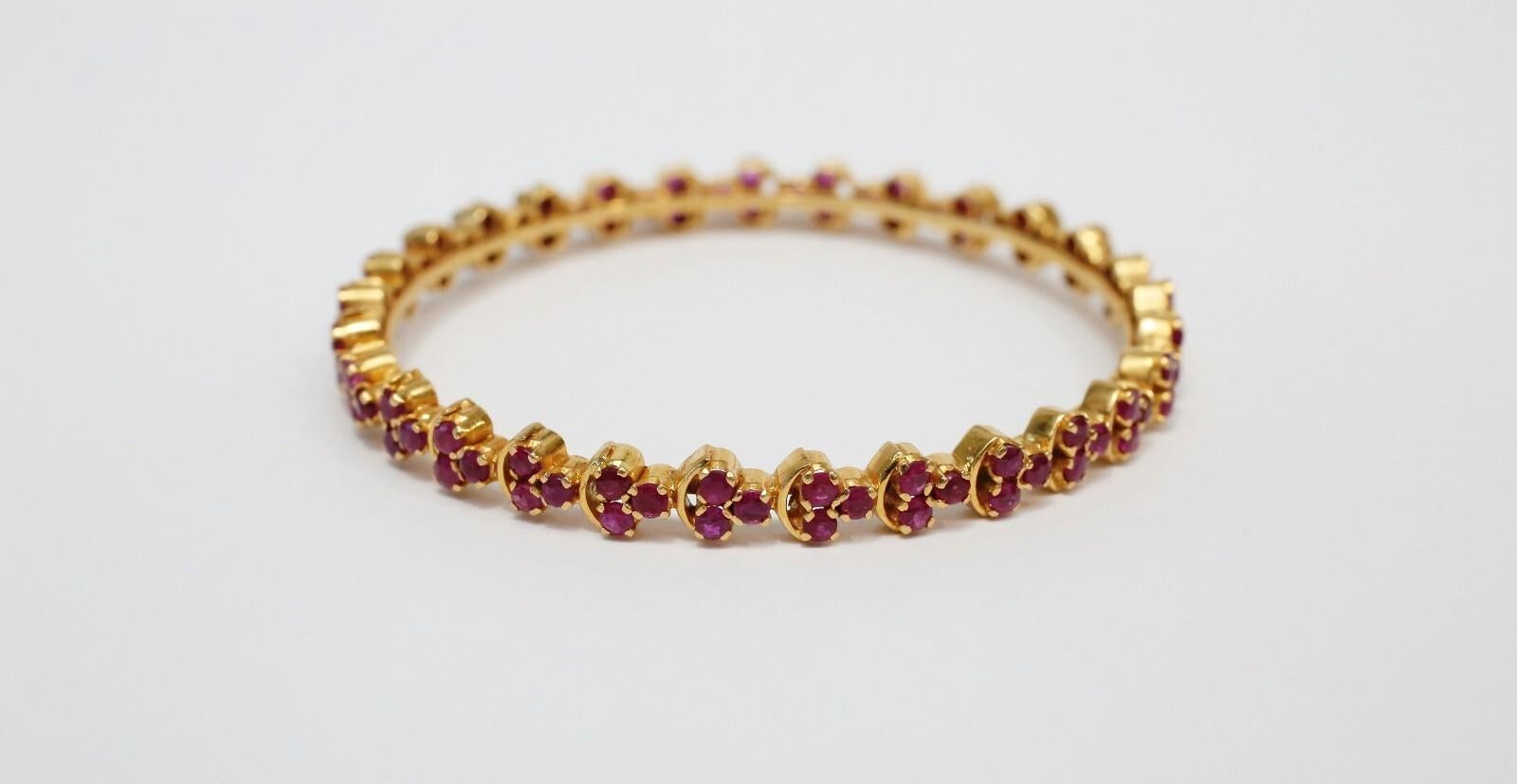 This elegant 21k yellow gold bangle showcases a striking array of vibrant rubies, each set in a repeating pattern of three-stone clusters. The gemstones are securely mounted in a scalloped channel setting that encircles the entire bangle, creating a