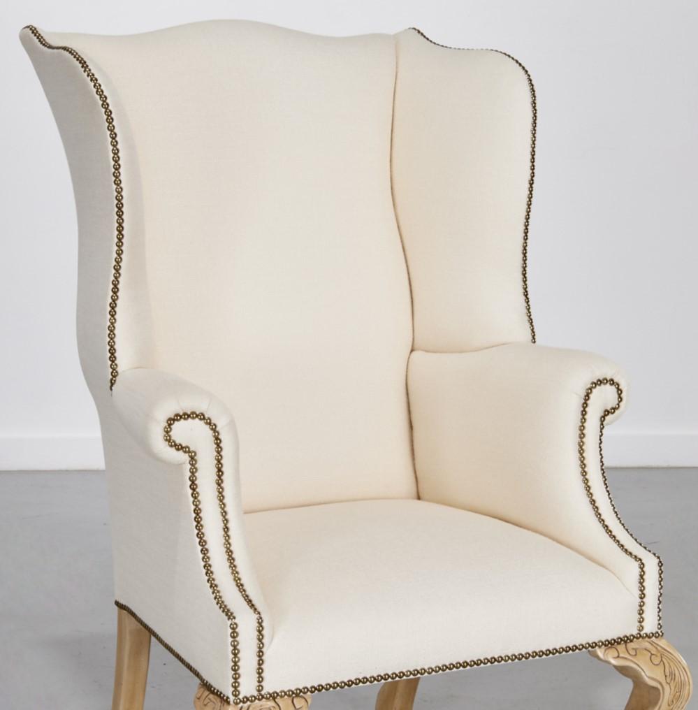 A.I.C., fauteuil à oreilles sur mesure de conception classique avec des touches modernes. Il est revêtu d'un tissu crème, souligné par des têtes de clous en laiton qui ajoutent un accent décoratif et traditionnel. Le dossier haut et les ailes