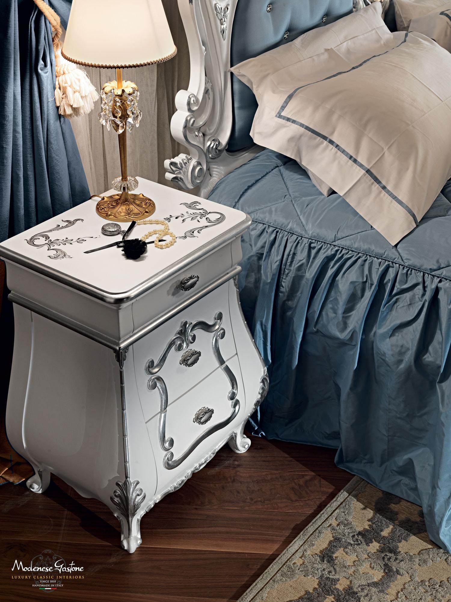 Elaborately carved baroque night stand by Modenese Luxury Interiors with white lacquered finish and silver leaf handmade decorations. This curved-shaped item, which is good in luxury bedrooms alongside beds, dressing tables and credenzas, features