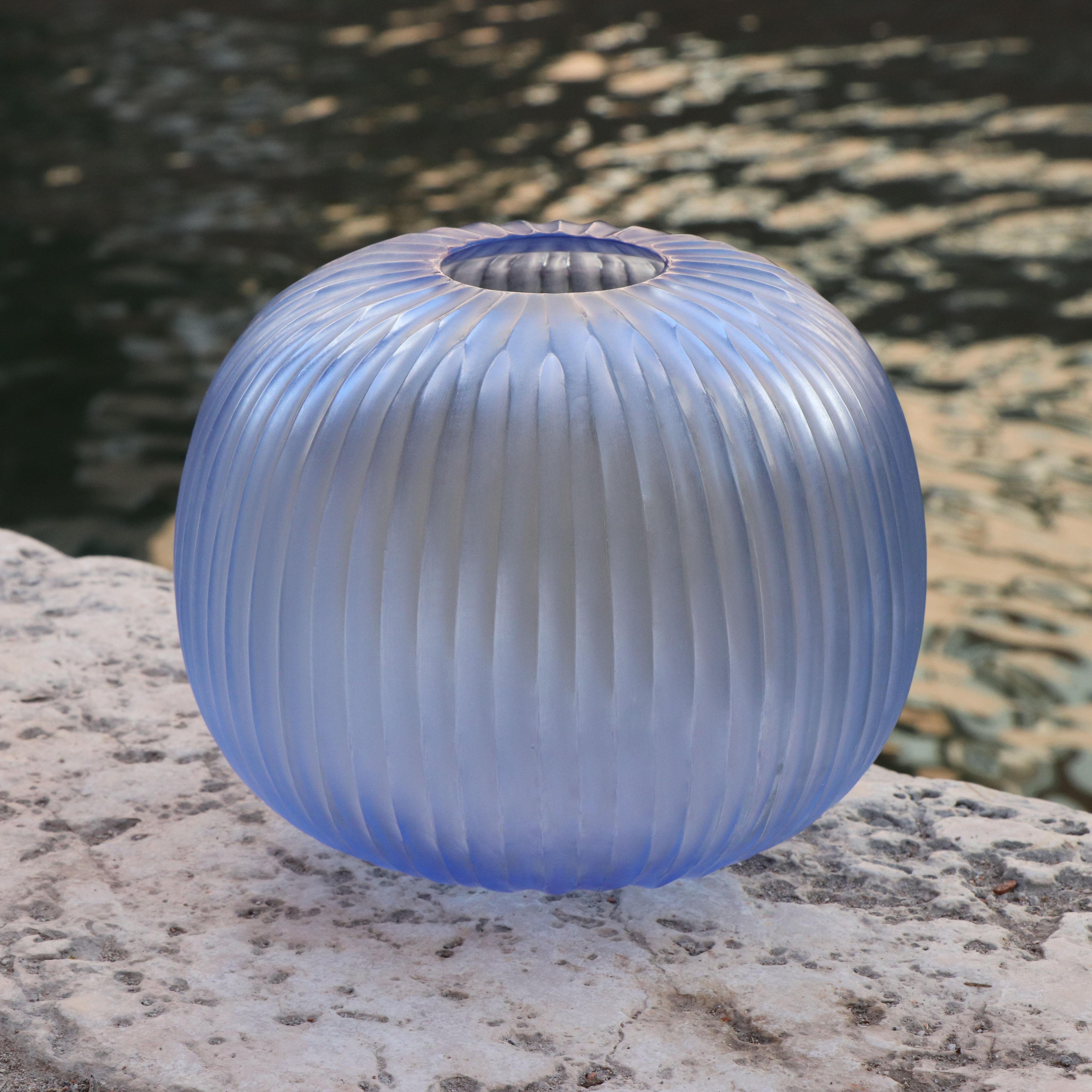 The melted glass is first blown in the heat of the furnace and shaped into a round vase with a small opening. Once solid the glass is cold carved to produce the linear cuts which mark its surface reminding us of the shell of a sea urchin –