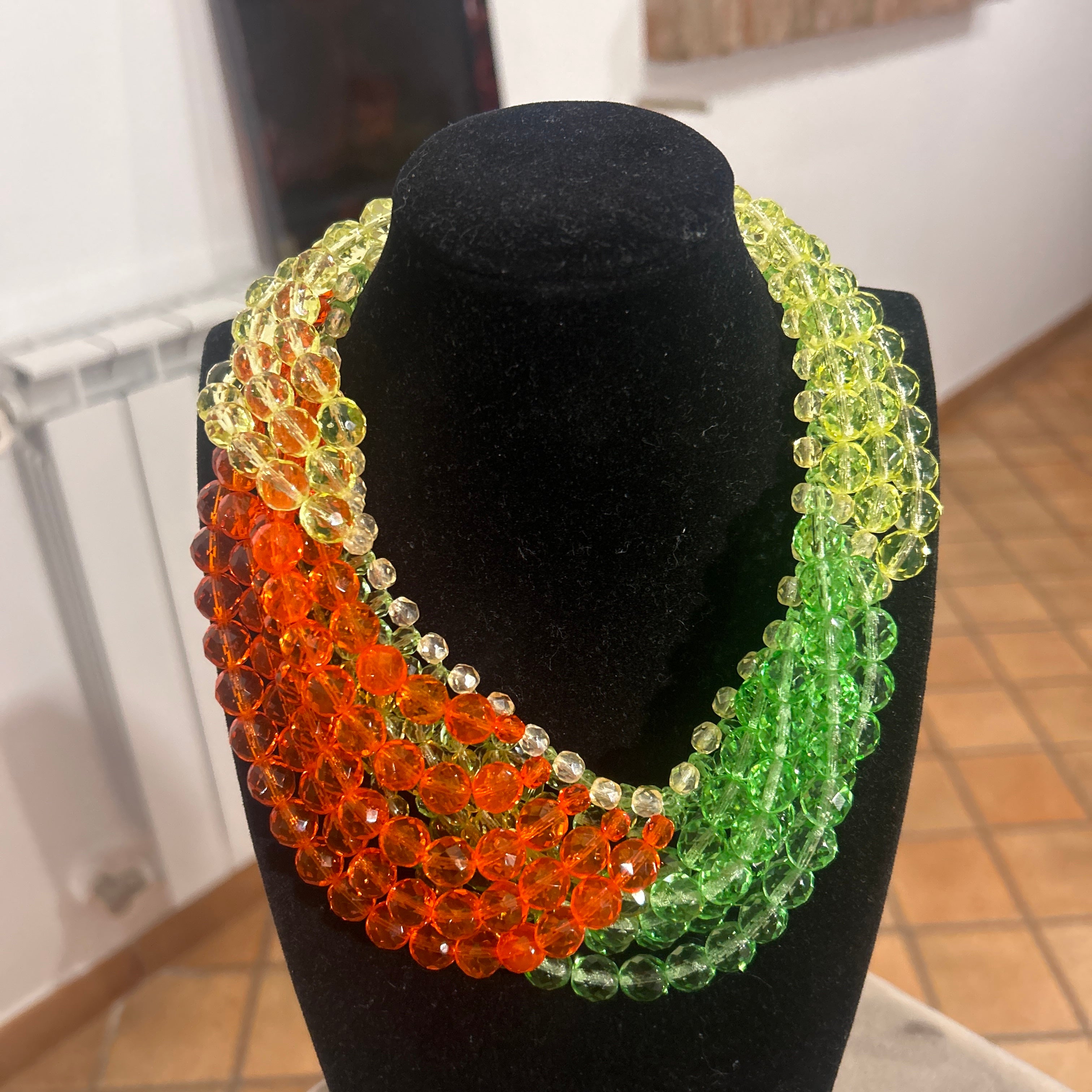 The necklace by Coppola e Toppo features a dramatic interplay of green and orange crystal beads, arranged in a richly layered, braided design. Multiple strands of faceted beads are intricately woven together, creating a bold, undulating pattern that