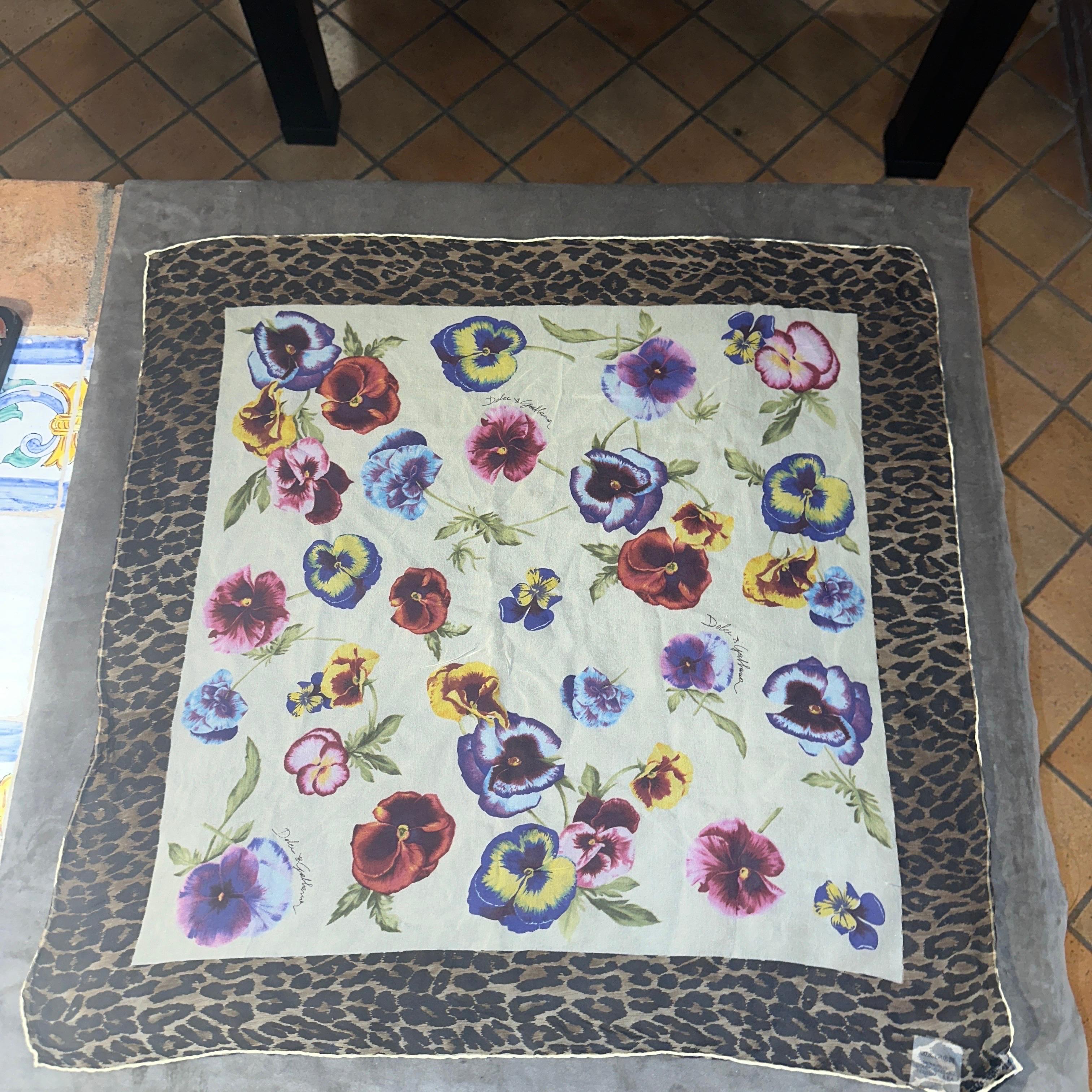 This is a Dolce & Gabbana scarf featuring a vibrant and eclectic design that blends natural and bold motifs. At the center, the scarf showcases a vivid floral pattern with pansies in rich hues of purple, yellow, red, and pink, scattered on a light