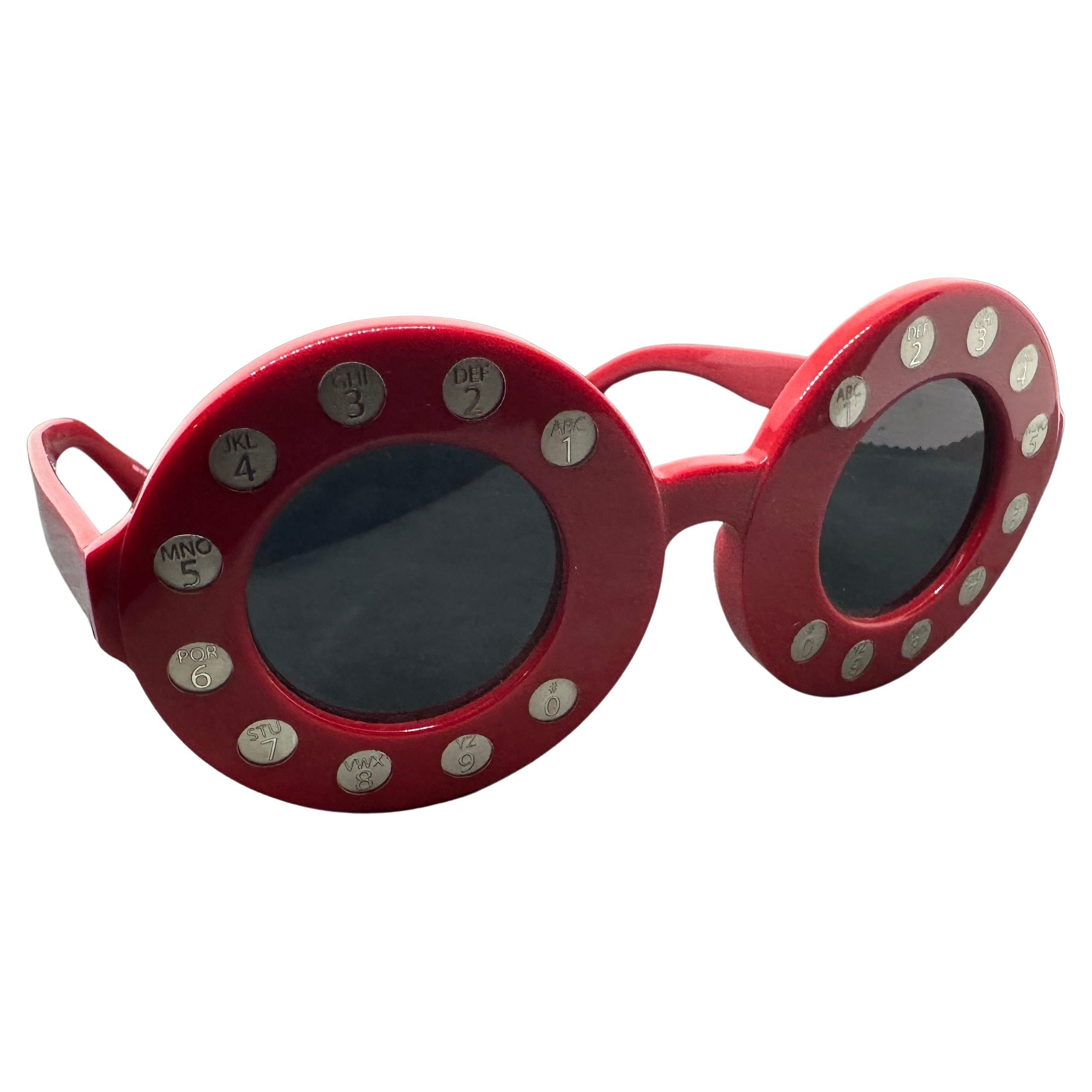 These sunglasses by Yazbukey feature a distinctive, bold design. They have a round shape with a bright red frame, accented by silver-toned numbers like an old telephone around the lenses. The lenses themselves are dark and offer a strong contrast