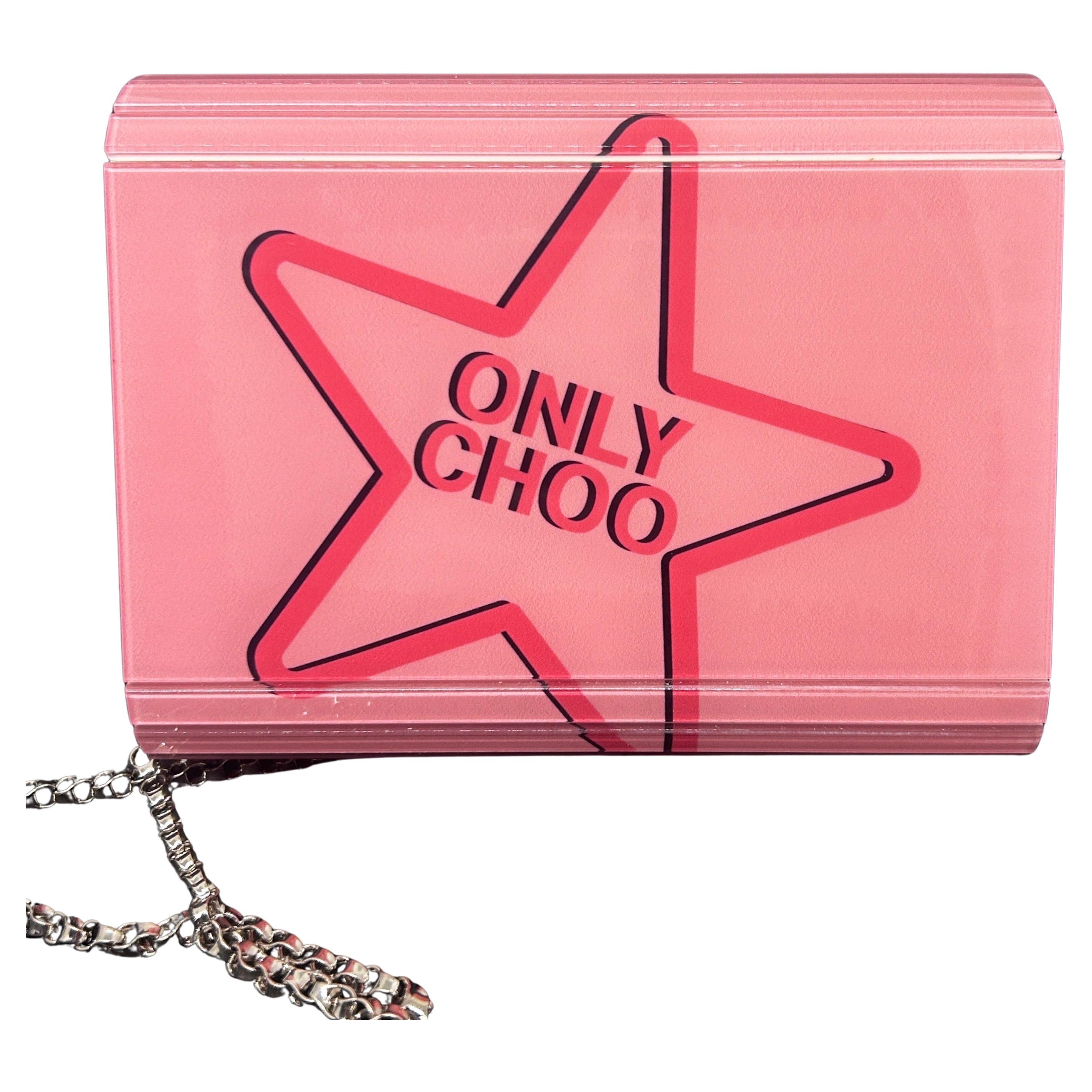 This Jimmy Choo clutch bag features a bold and playful design with a hard, structured pink acrylic shell. On the front, there’s a large red heart graphic outlined in black, with the words 