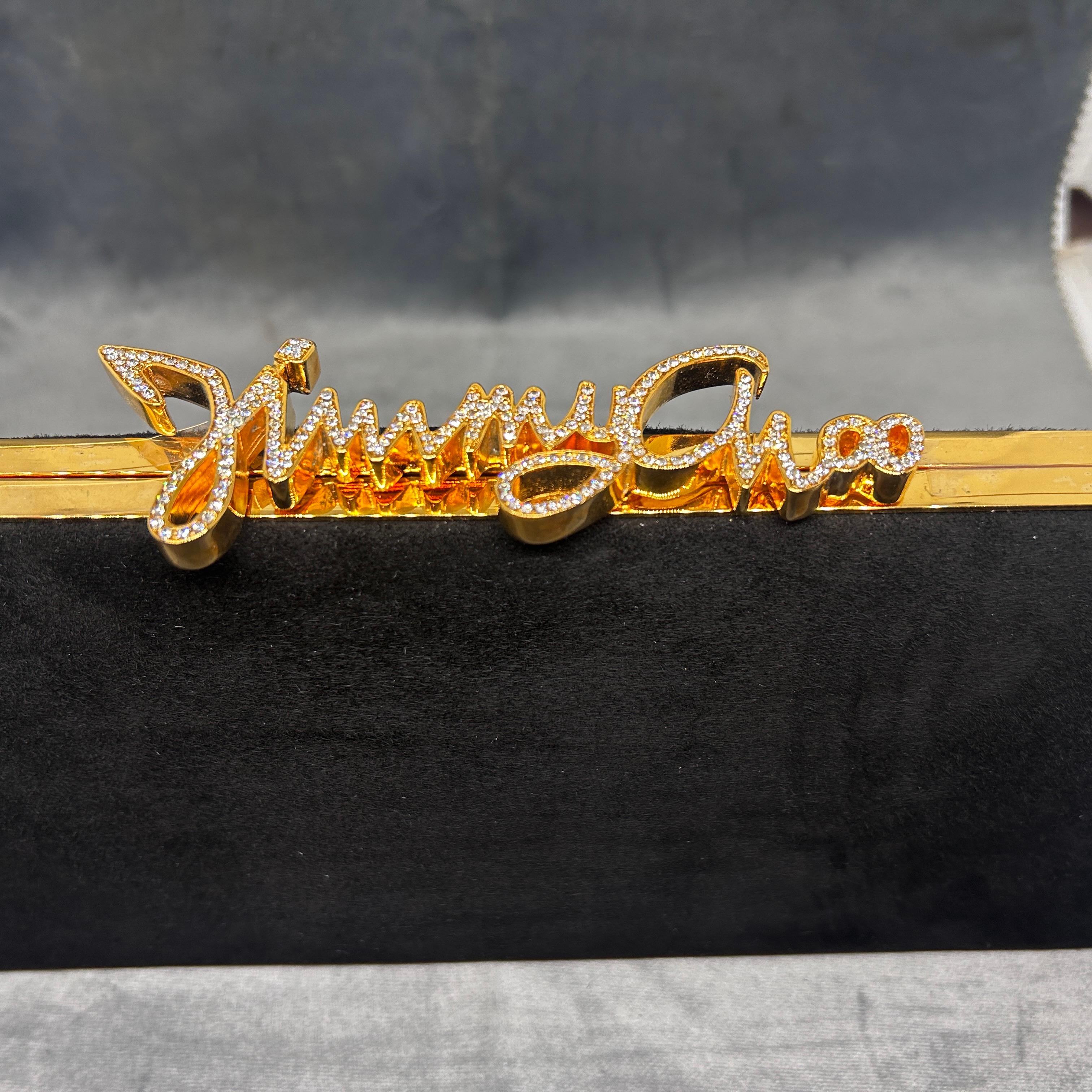 This Jimmy Choo clutch bag features a sleek and elegant design. The body of the clutch is rectangular with a black exterior, likely made from a smooth or velvet-like material that gives it a refined and luxurious appearance. The top edge of the