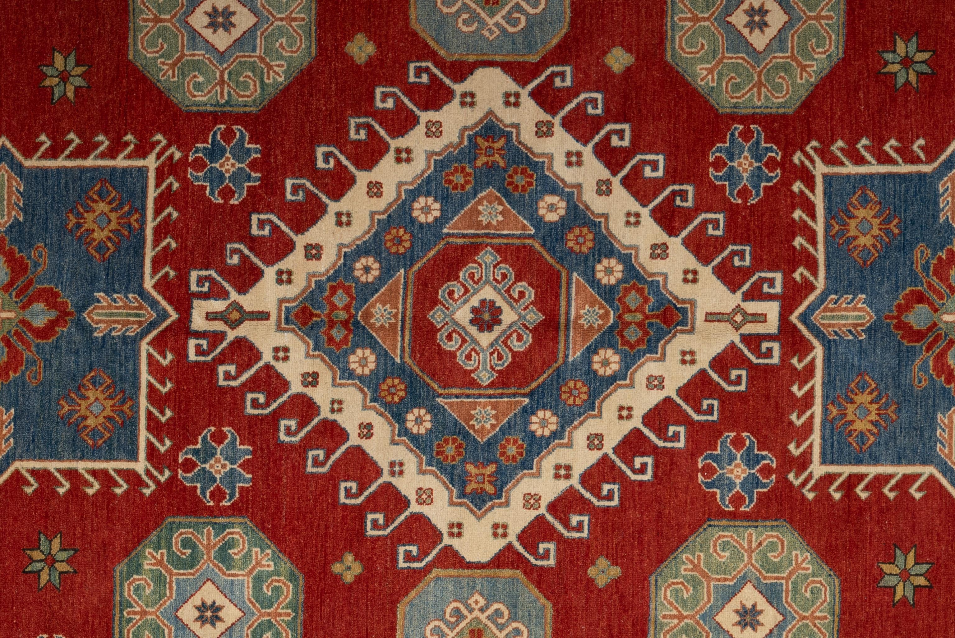 Esta vibrante alfombra muestra una audaz geometría y un rico contraste de colores en una llamativa paleta de rojo rubí, azul vaquero y topo. Anudada a mano en lana, presenta poderosos motivos tribales -como medallones en forma de gancho, estrellas