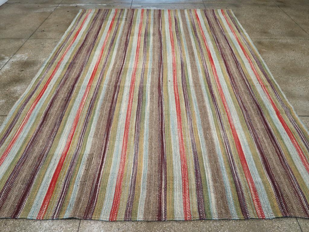 Esta alfombra persa Flatweave Kilim del siglo XXI, que mide 3,8 m x 4,8 m, muestra una disposición vibrante y rítmica de rayas verticales en una gama de tonos terrosos y cálidos. La composición presenta una armoniosa mezcla de azules apagados,