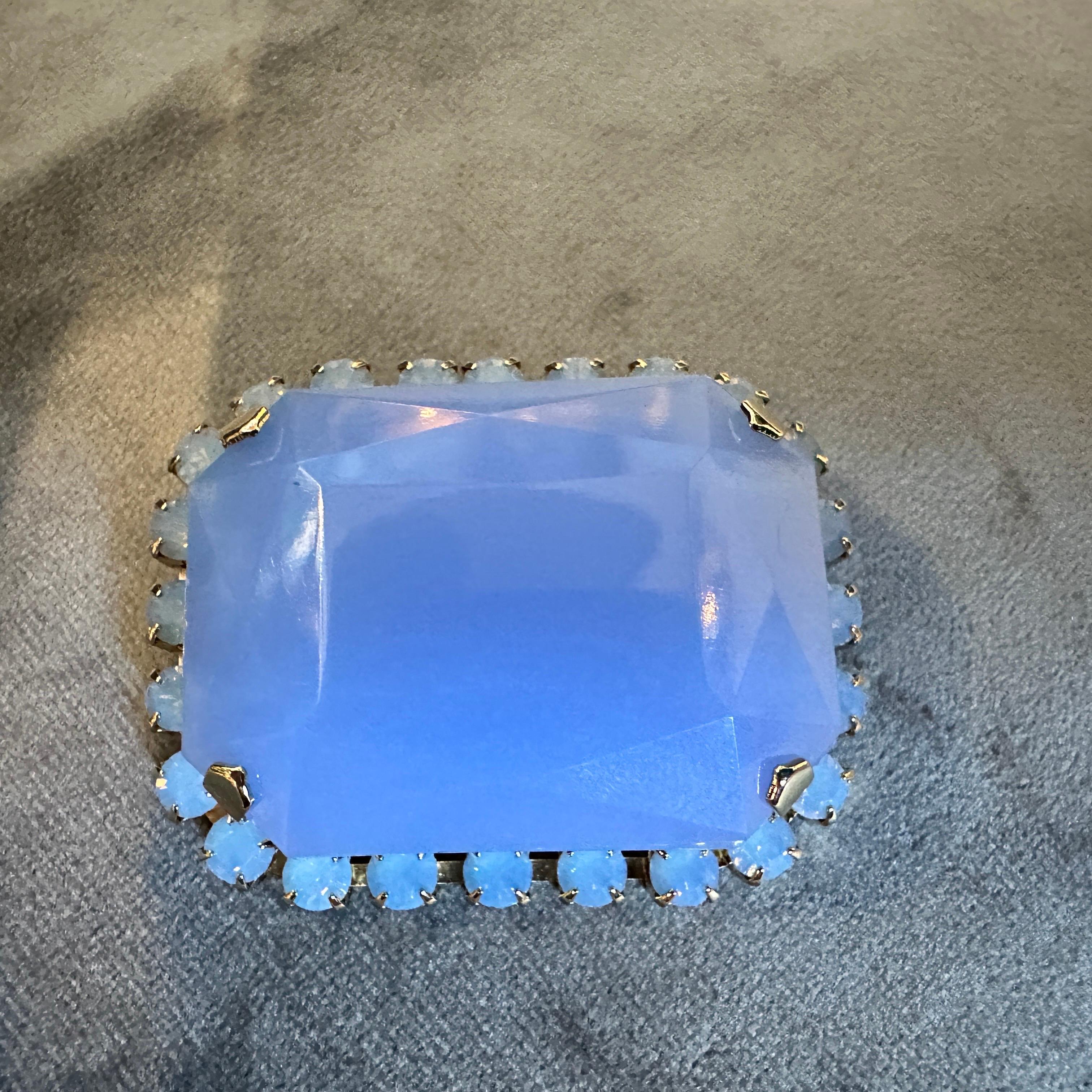 Questa spilla del XXI secolo, mai usata, di Coppola & Toppo, è caratterizzata da un'estetica retrò grazie all'uso di resina blu opalescente e metallo dorato. Al centro c'è una grande pietra di resina sfaccettata in una morbida e luminosa tonalità
