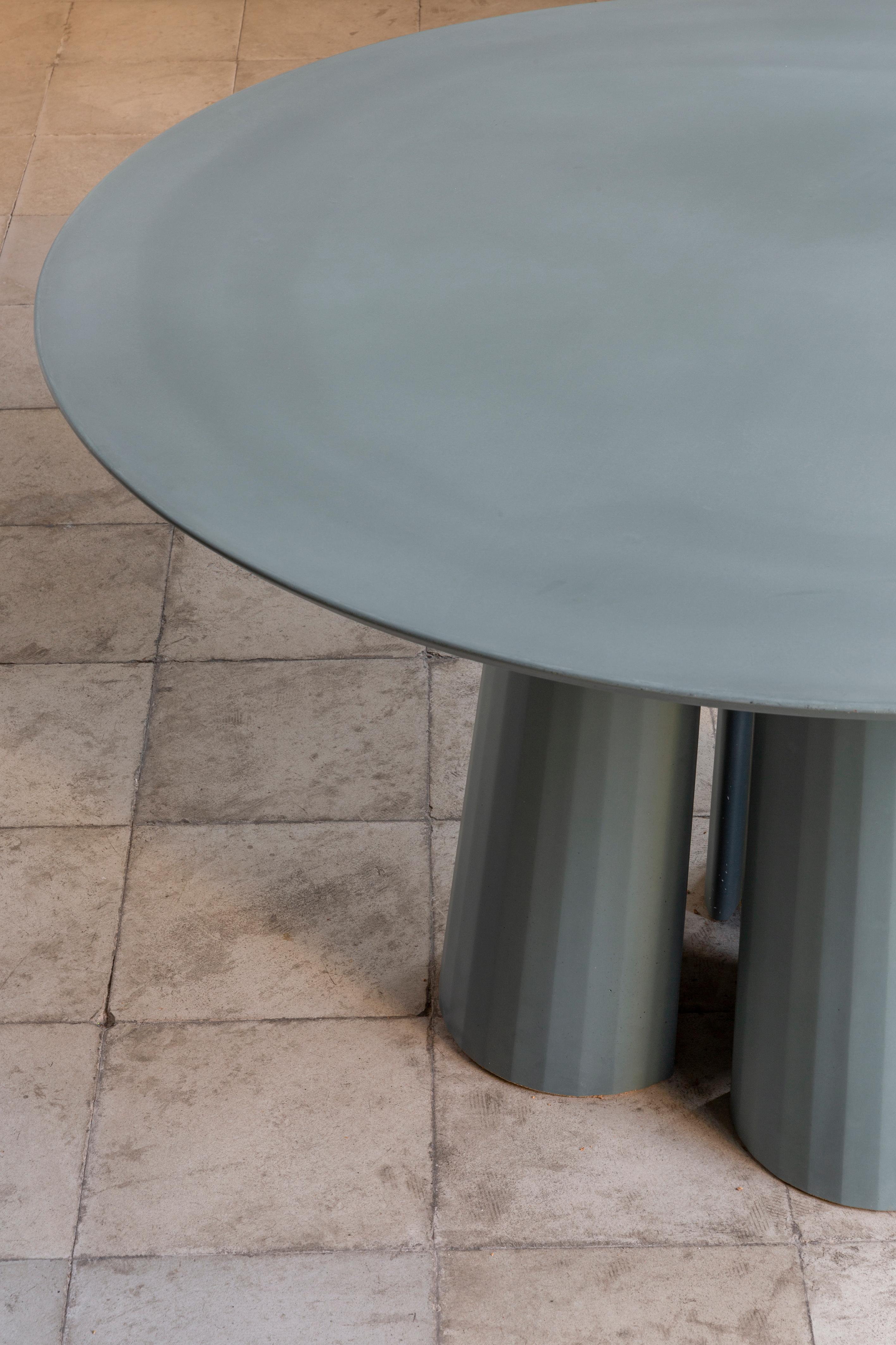 Rounded shape dining table part of a collection of modular system in ultra-high performing cement mortar. UHPC tabletop and base colored in the mixture and sandblasted. Available in eight different colors: Brick, silver, powder, cream, fir, dark,