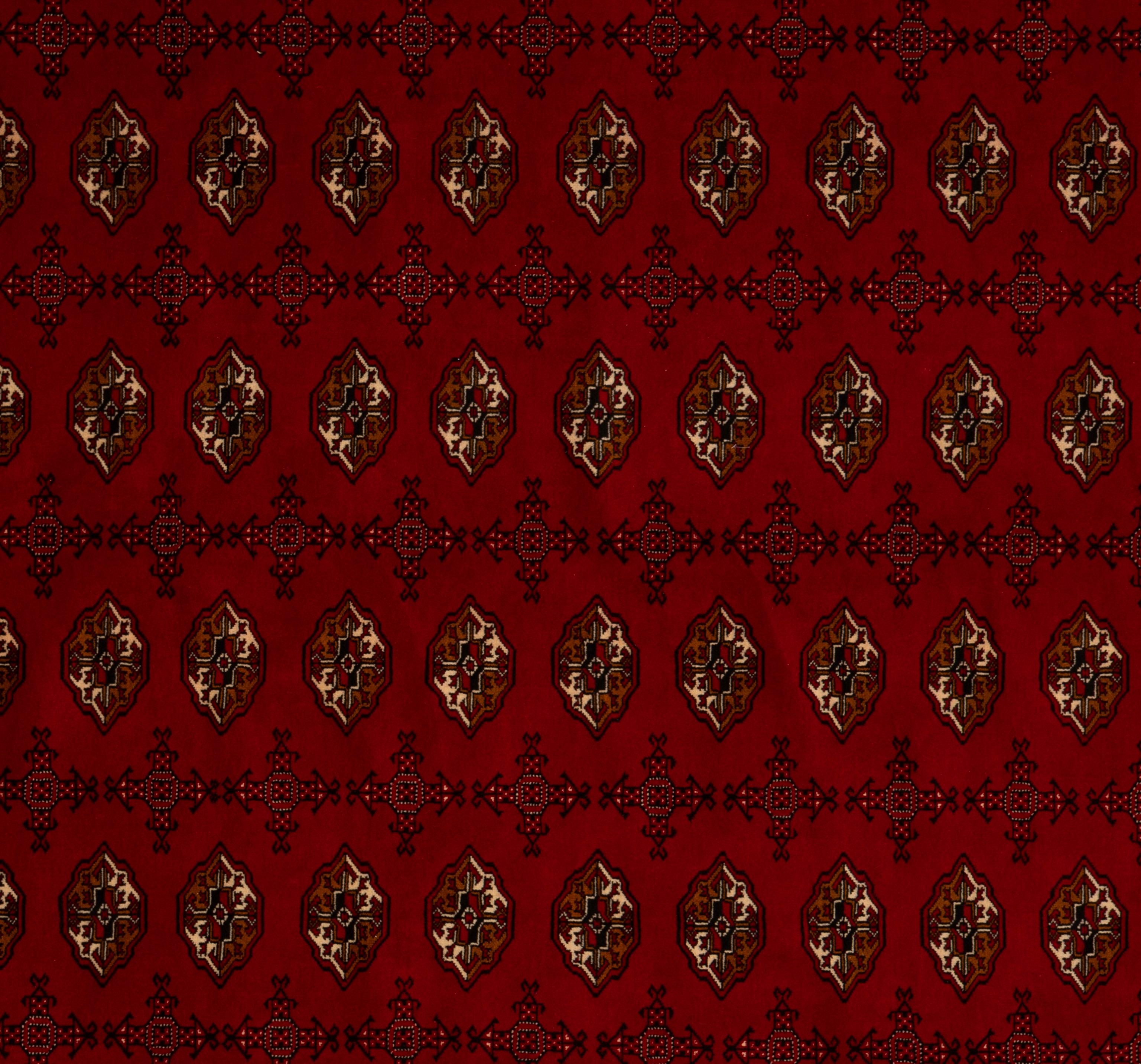 Esta alfombra tribal Bokara está anudada a mano con lana fina y presenta un rico fondo rojo rubí que resalta maravillosamente sus icónicos motivos gul repetidos. El profundo campo de tonos joya está enmarcado por múltiples bordes de intrincados