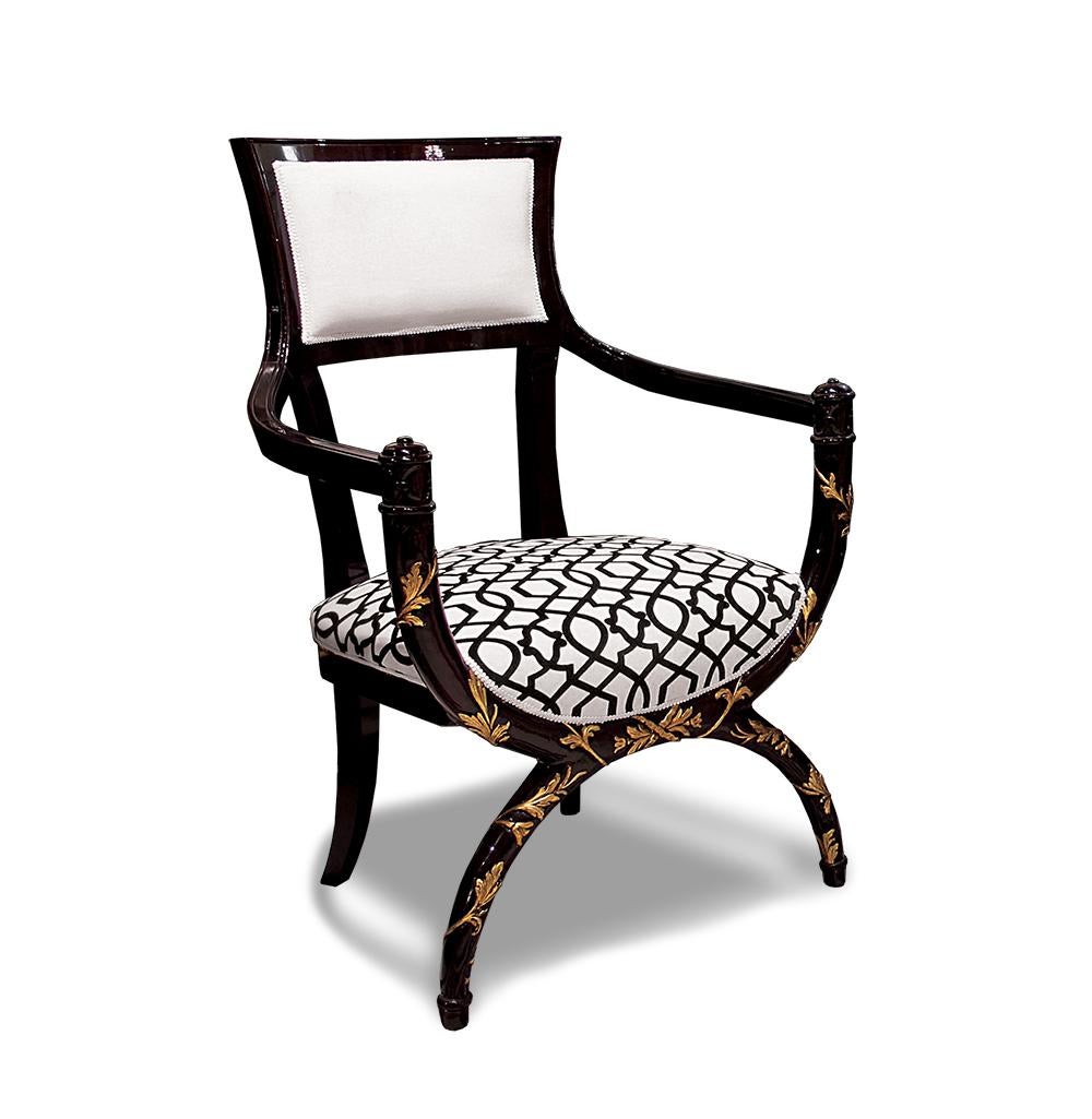 This elegant empire style armchair is inspired by the glamour of the 30s. Its sinuous structure is in mahogany, finely adorned with carved decorations highlighted with gold leaf. The cushions are covered in precious velvet and the seat features an