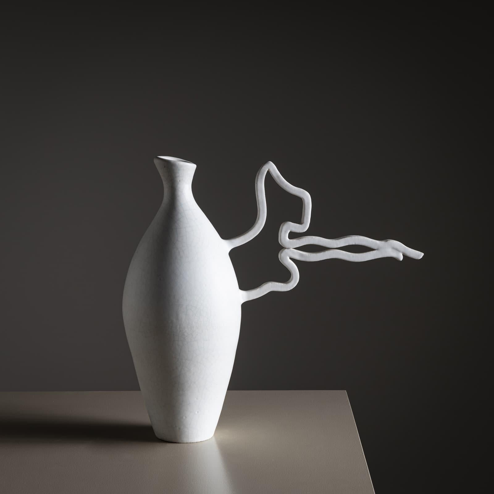 

Vase en céramique émaillée blanche avec un corps conique lisse et une longue anse de forme libre sur le côté. La surface présente une fine craquelure. Le vase est signé POEMU / de Mons / Aix sur le fond.

Poemu est un artiste français
