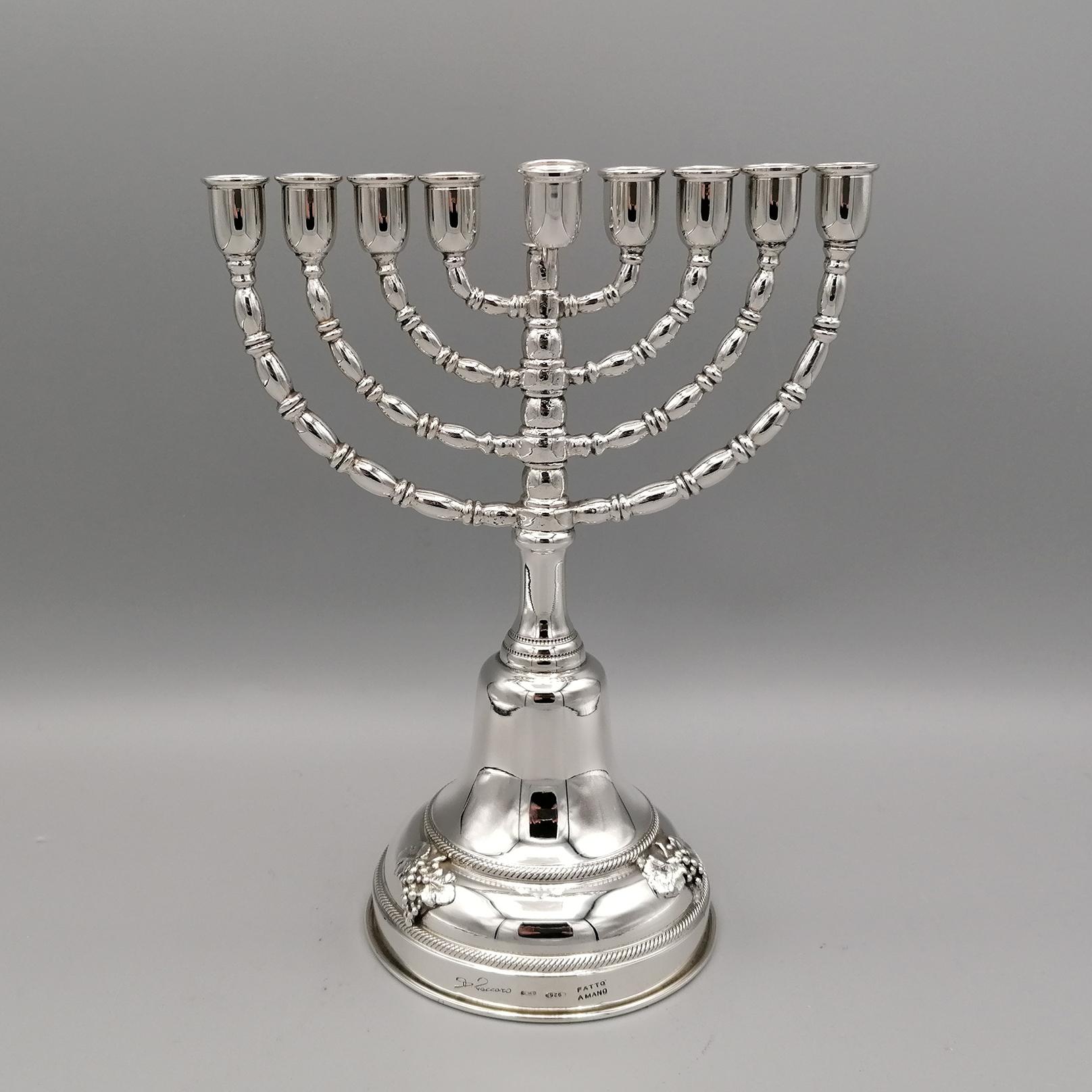 Hanukkah in sterling silver. The base is round shaped. Between two frames with a rope motif, 3 bunches of grapes with branches and leaves were applied for decoration. The central stem is shaped while the 8 arms are made with oval and ring motifs.