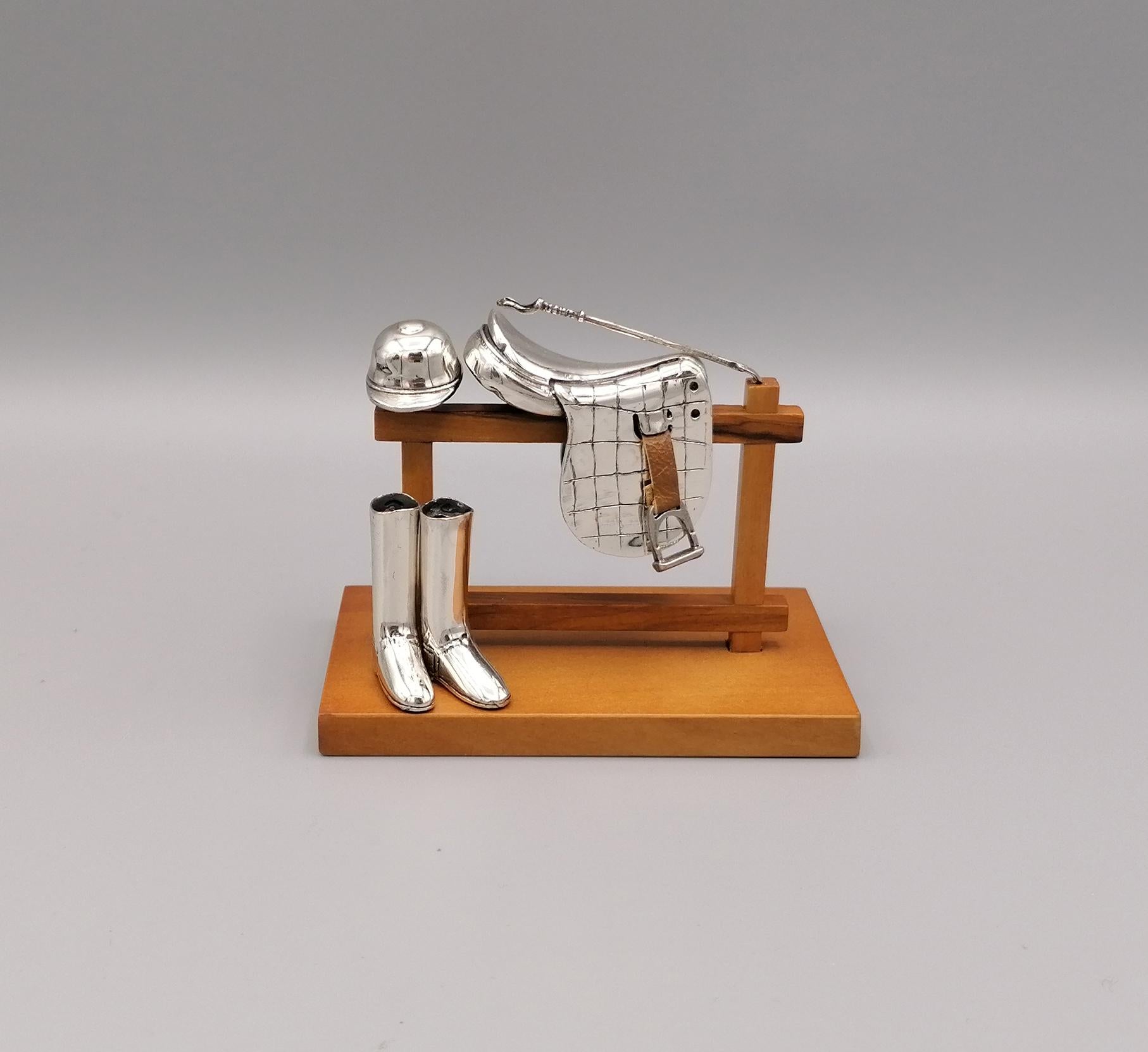 Miniature in 925 sterling silver depicting a saddle, boots, hat and whip for a jockey resting on a wooden fence.
The workmanship is in fusion and finished with chisel.
The object can be customized on request with a silver plaque, max size cm. 2 x