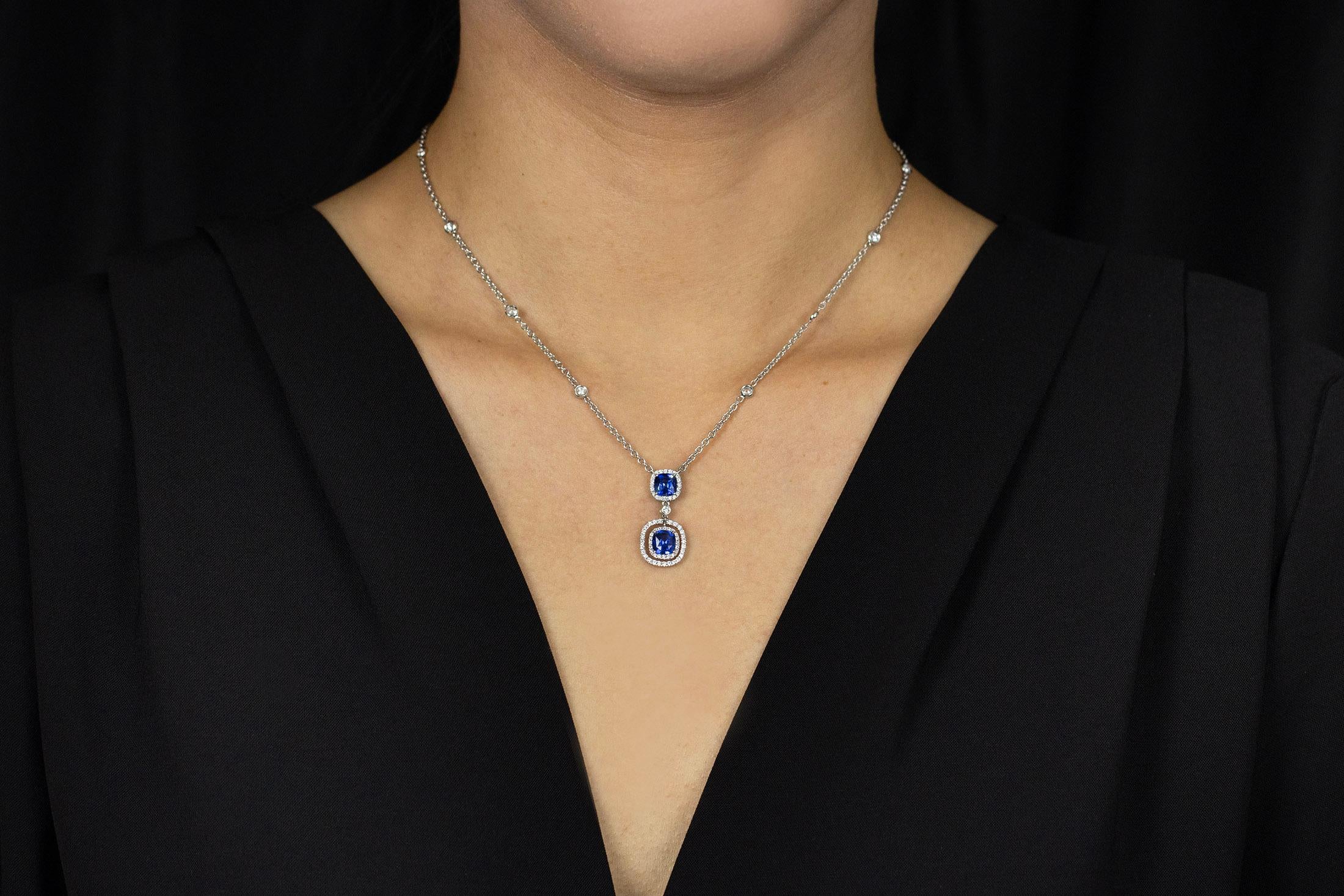 An elegant pendant necklace showcasing two cushion cut blue sapphire weighing 2.20 carats total. Each blue sapphire is surrounded by a row of brilliant round diamonds in a halo design weighing 0.75 carats total, and a bezel set round diamond spaced