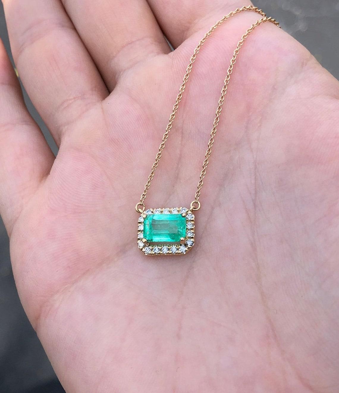 Featured here is a stunning East-to-West Colombian emerald and diamond halo necklace made in fine 14K yellow gold. Displayed is a lustrous, medium-light green with incredible transparency, accented by a simple four-prong 14K mount, allowing for the