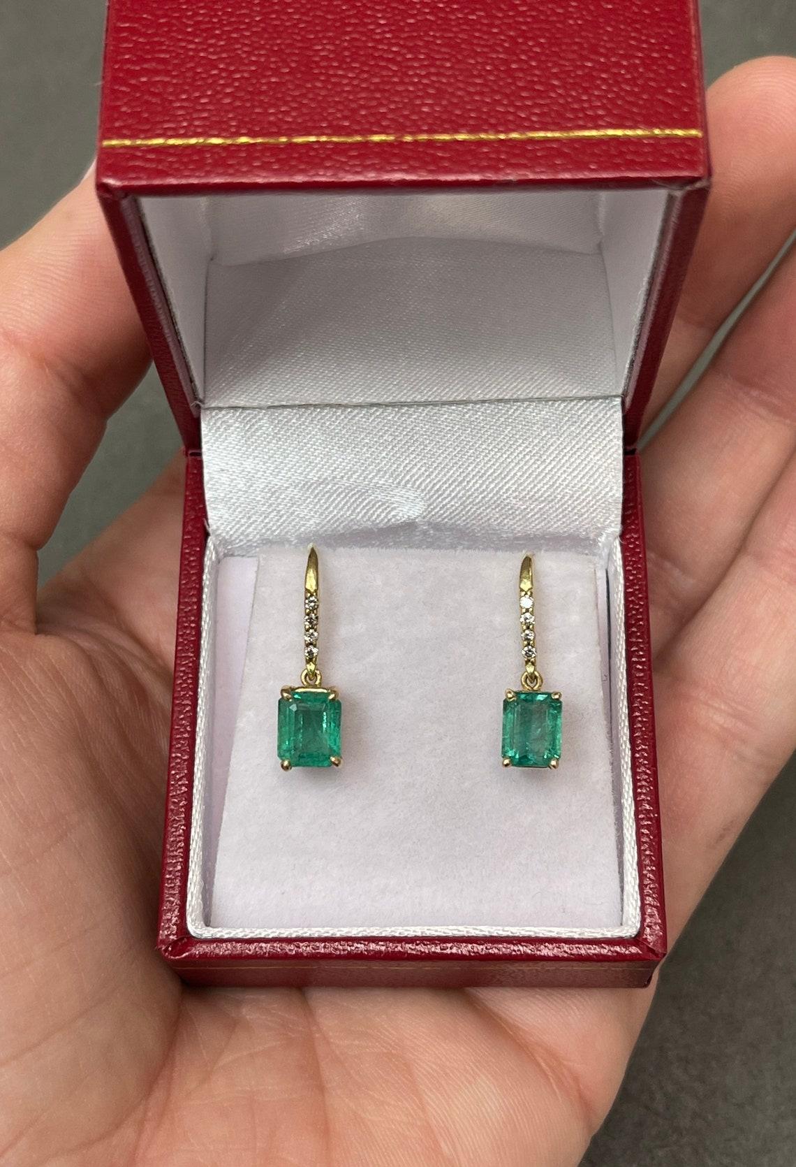 A stylish pair of emerald cut-emerald & diamond hook earrings made in 18K yellow gold. The chic pair of earrings feature green, natural emeralds. The emeralds have beautiful eye clarity and are simply vivacious! The gems display minor flaws that are
