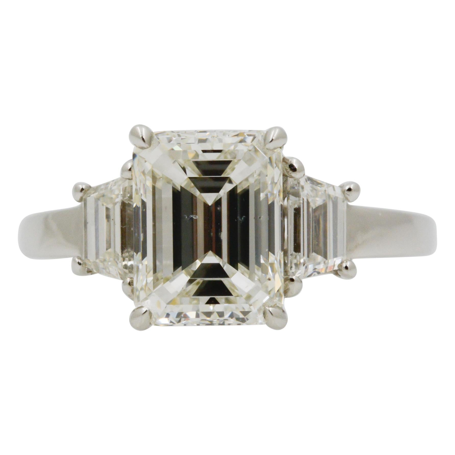 This platinum three stone diamond ring features a 2.29 carat emerald cut GIA certified diamond with I coloring and S12 clarity. The center stone is surrounded by two trapezoid diamonds (0.74 carats combined, I color, and VS2/SI1 clarity). The ring