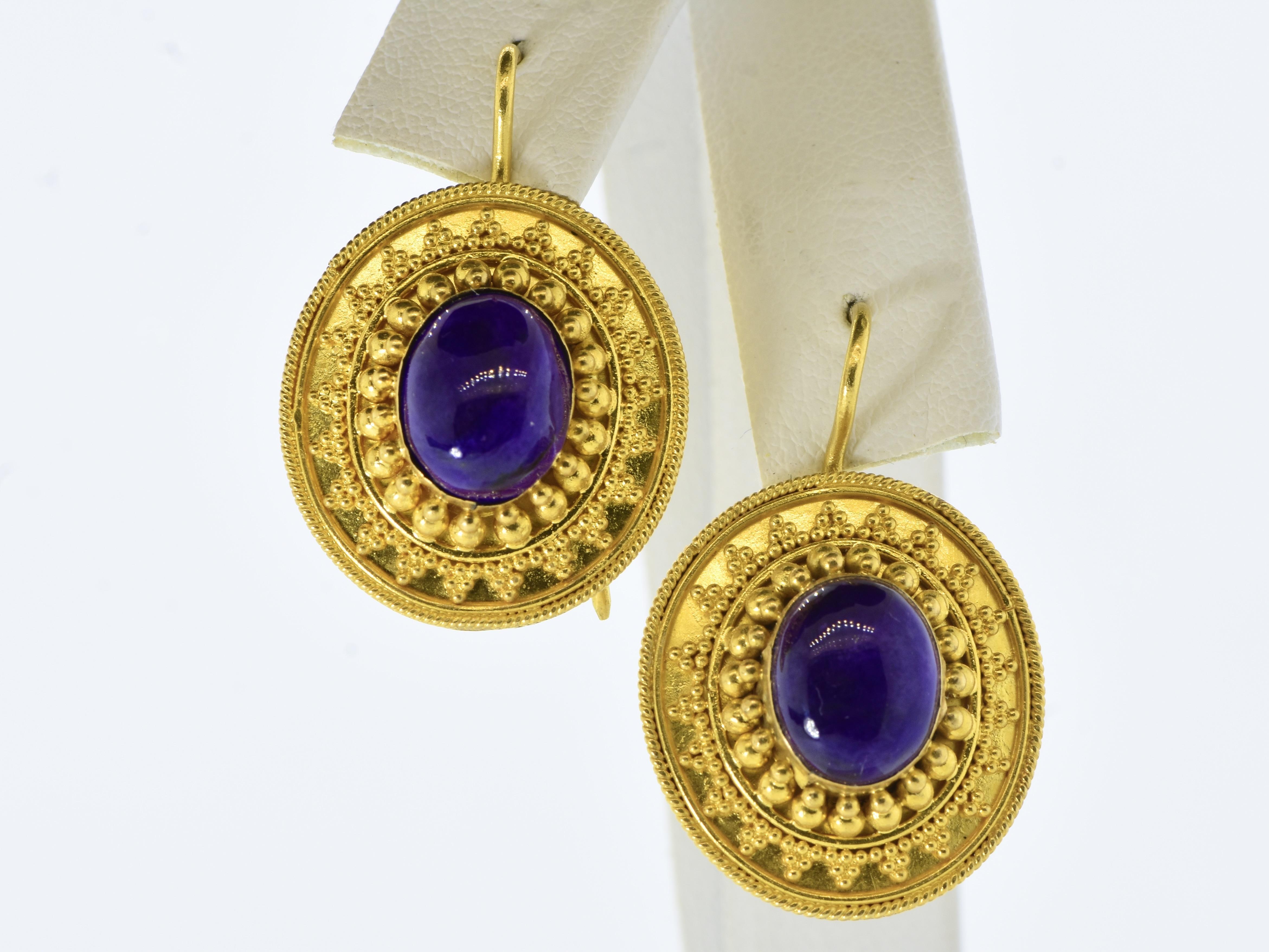 22k gold earrings with intricate detailing. These contemporary earrings feature a bold design with a central deep purple cabochon stone. The gold is crafted into an ornate pattern with raised bead-like accents and rope motifs surrounding the stone.