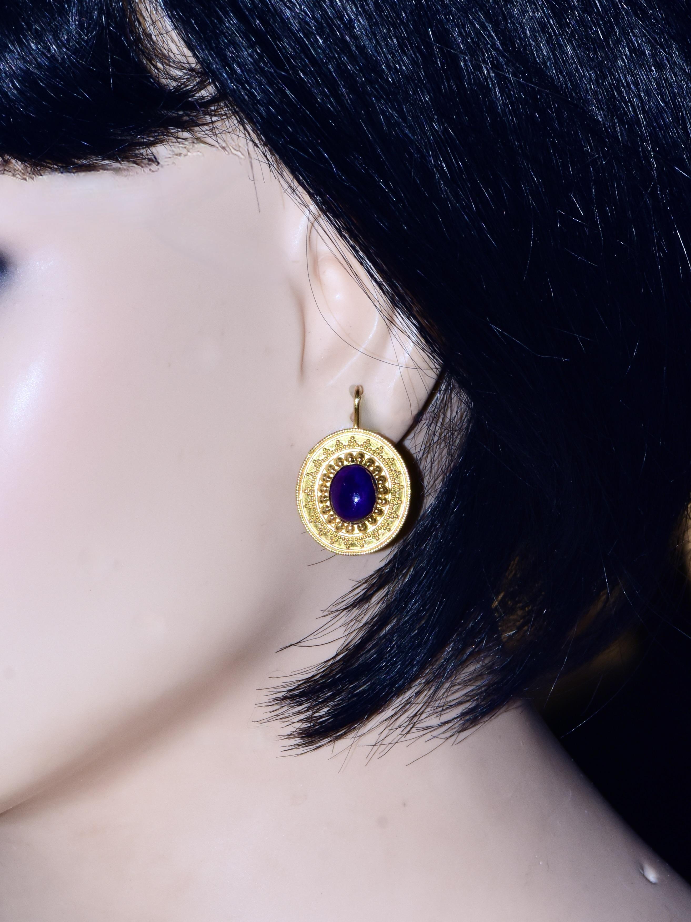 Orecchini in oro 22k con dettagli intricati. Questi orecchini contemporanei presentano un design audace con una pietra cabochon centrale di colore viola intenso. L'oro è lavorato in un motivo ornamentale con accenti in rilievo simili a perline e