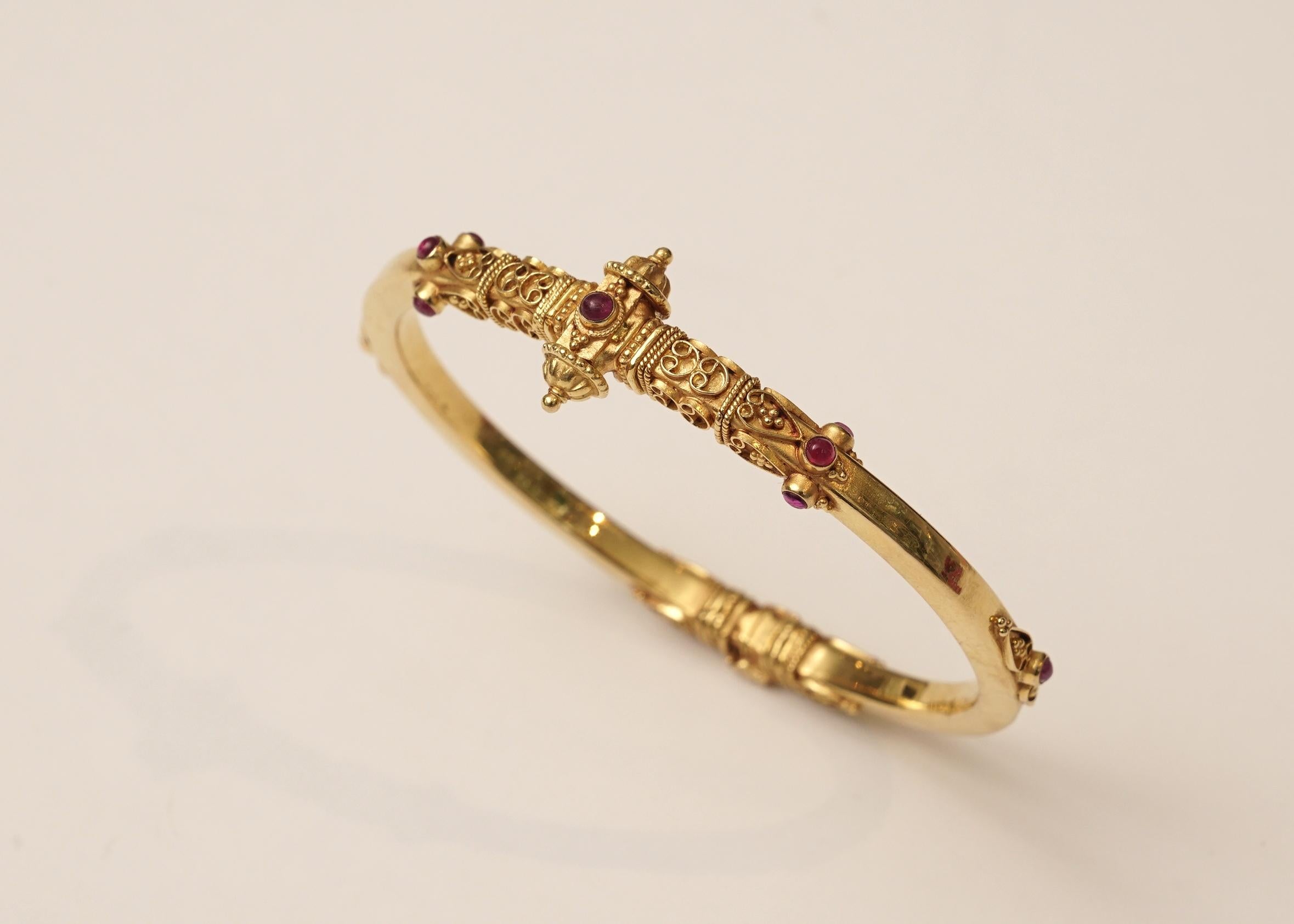 A 22K gold bangle with intricate granulation and wire work on the top, sides and bottom and set with cabochon rubies.  Hinged at the bottom, and one side of the top unscrews to open to release the clasp.  The screw has a stop so that it will not