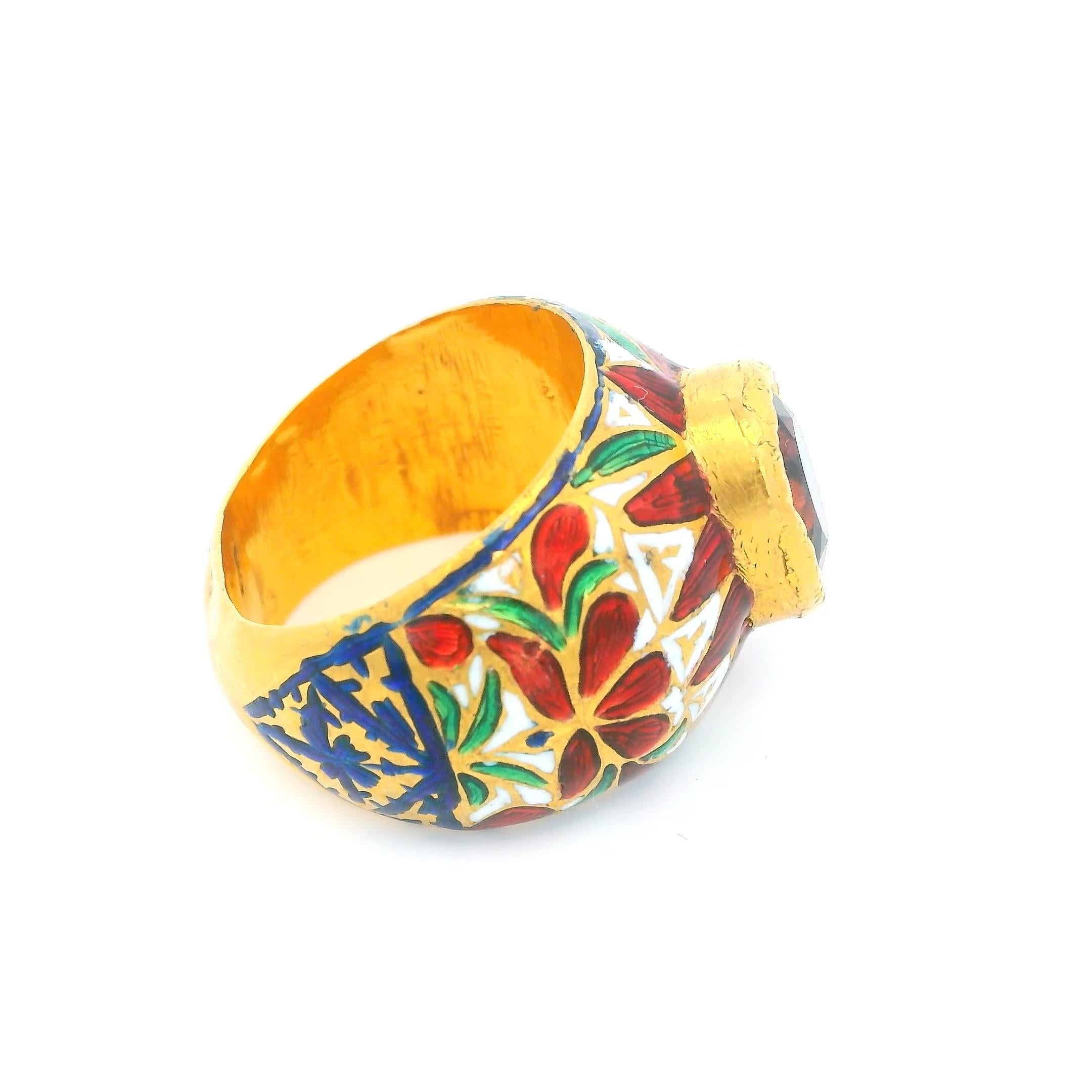 A vibrant and ornate ring featuring a striking oval gemstone in a deep red hue.

The band is adorned with intricate enamel work, showcasing red and green floral motifs that encircle the gemstone.

Crafted in 22k gold, this exquisite piece reflects