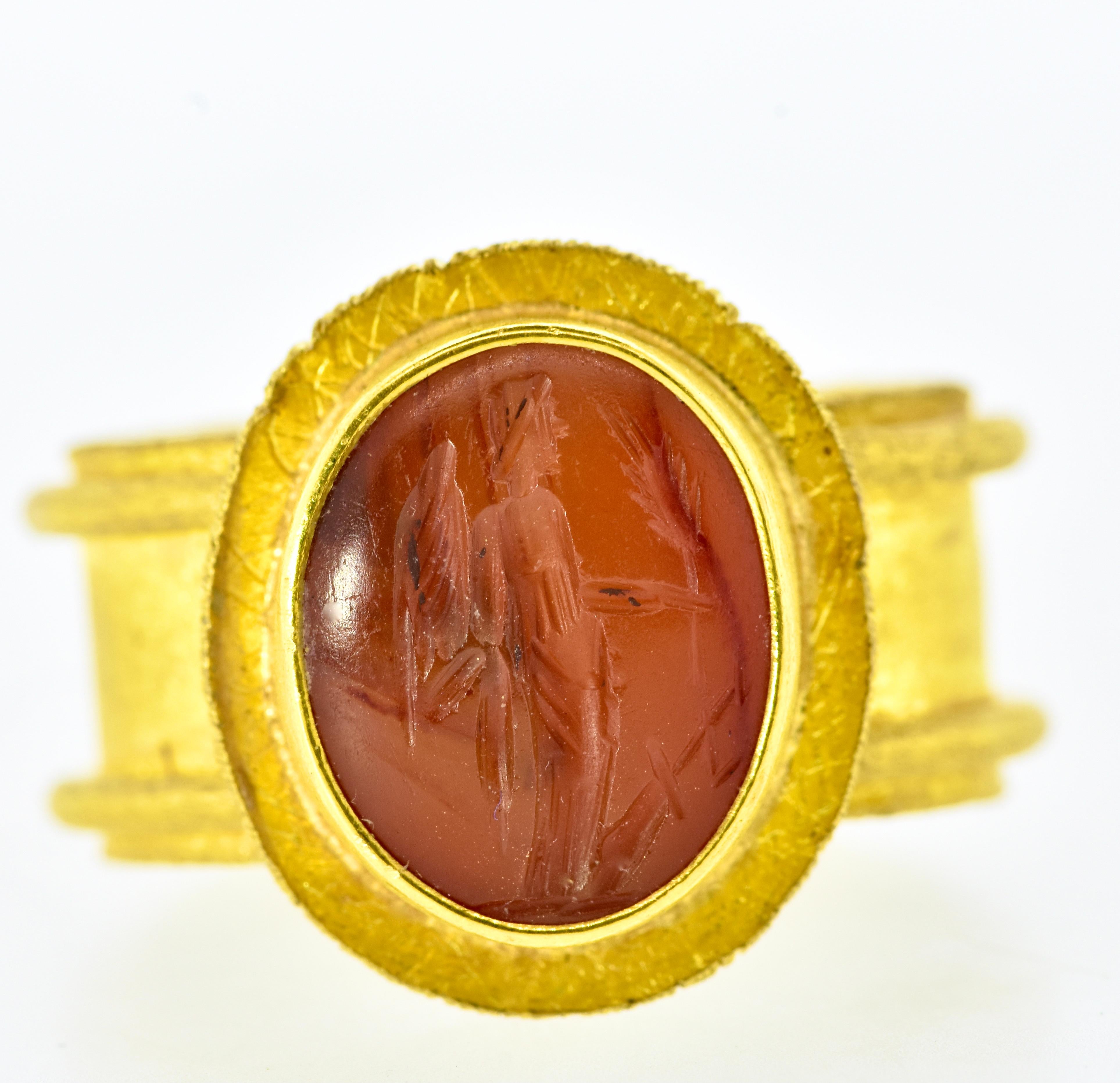 22K yellow gold ring centering an antique carnelian intaglio of a standing angel extending an olive branch - signifying peace. The intaglio measures 14.4 mm by 11.5 mm. The wide band measures 10.1 mm.  This ring is signed by the jewelry designer