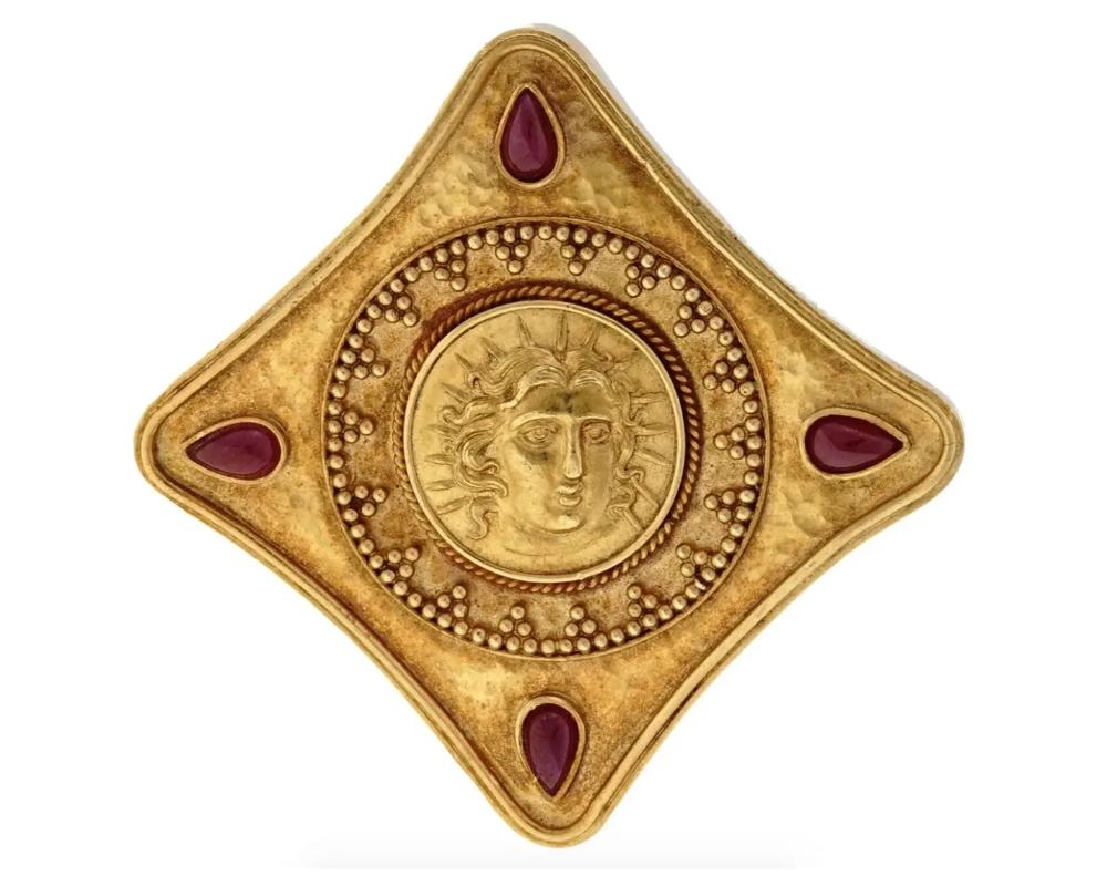 A vintage 22K yellow gold brooch pendant. The rhombus-shaped piece is garnished with an embossed portrait of the Ancient Greek god Apollo. The portrait is encompassed with a granulated ornament. Four ruby cabochons of teardrop shape are set on the