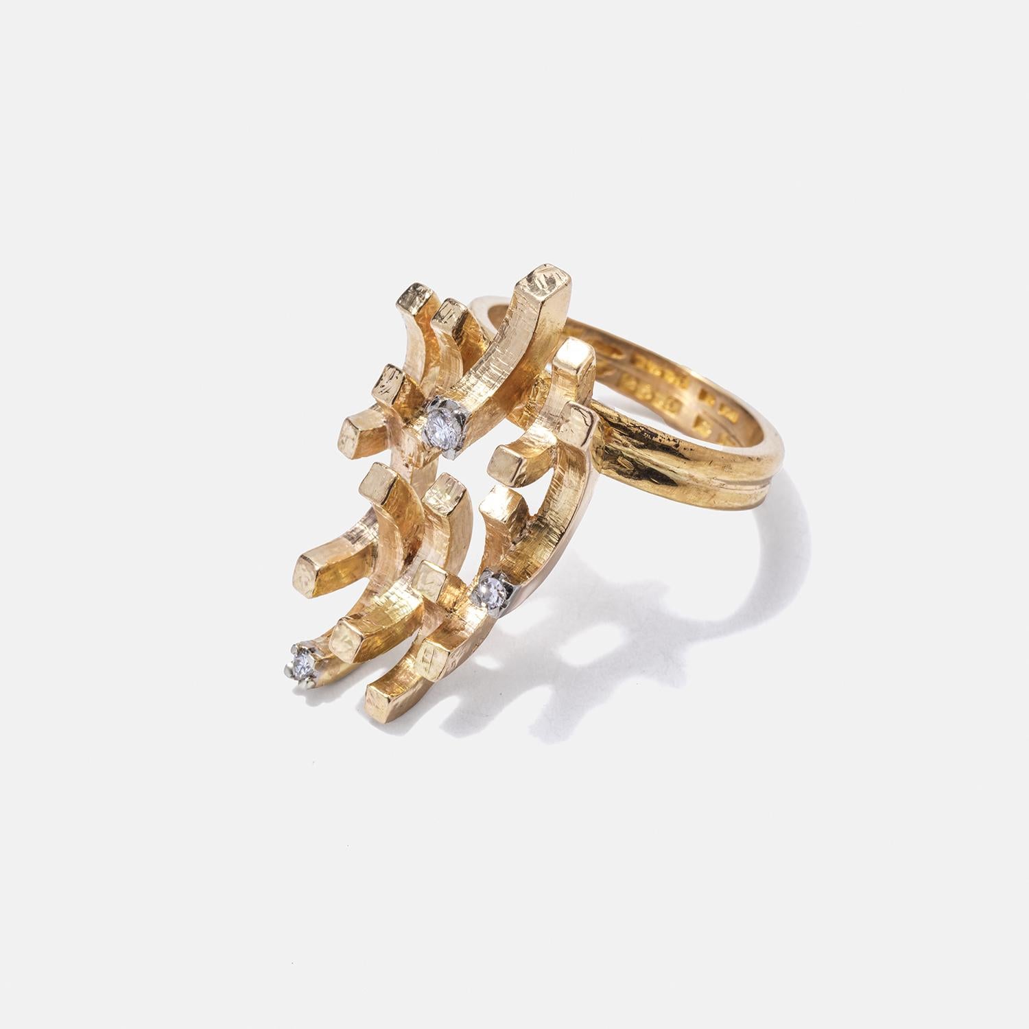 Der Schaft dieses exquisiten Rings besteht aus zwei 23-karätigen Goldbändern, die miteinander verschmolzen sind und so eine robuste und elegante Basis bilden. Darüber befindet sich ein Ornament aus 18-karätigem Gold, das einer kleinen, zarten Krone