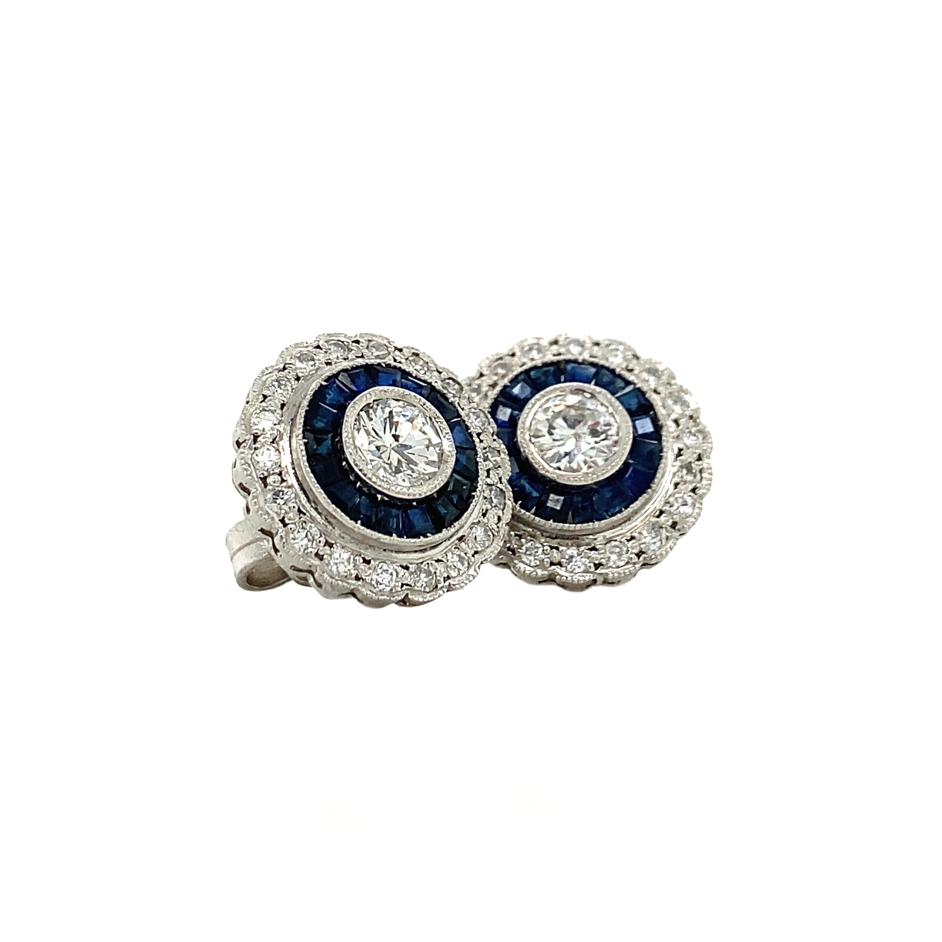 Uno squisito paio di orecchini a perno con un diamante da 0,25 carati incastonato in ciascuno, con un'aureola di altri 1,05 carati di diamanti, ulteriormente circondati da zaffiri taglio calibro. Realizzati in prezioso platino, questi orecchini