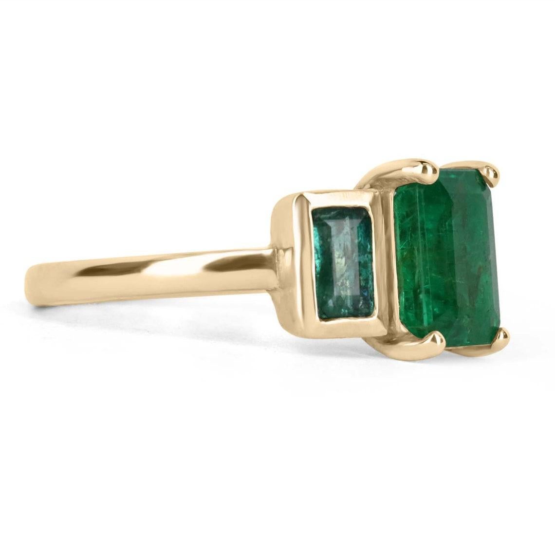 A Zambian emerald three-stone trilogy ring. This bespoke ring design is handcrafted in gleaming 14K solid yellow gold. The gorgeous piece features a rich dark green four-prong 1.75-carat emerald cut in its center. Emerald cut emeralds are flanked on