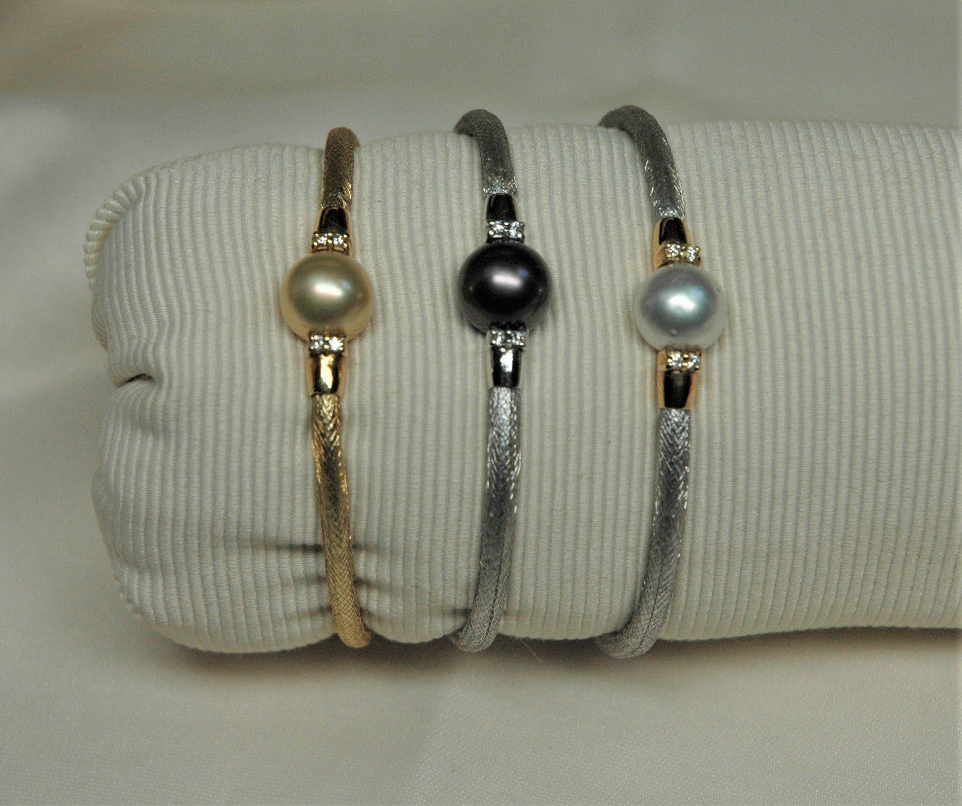 Three semi-rigid bracelets made of yellow and white gold with pearls and diamonds. The bracelet is made with an intertwining system that covers an internal structure. Each bracelet mounts a pearl of a different color: white, gold, and black.