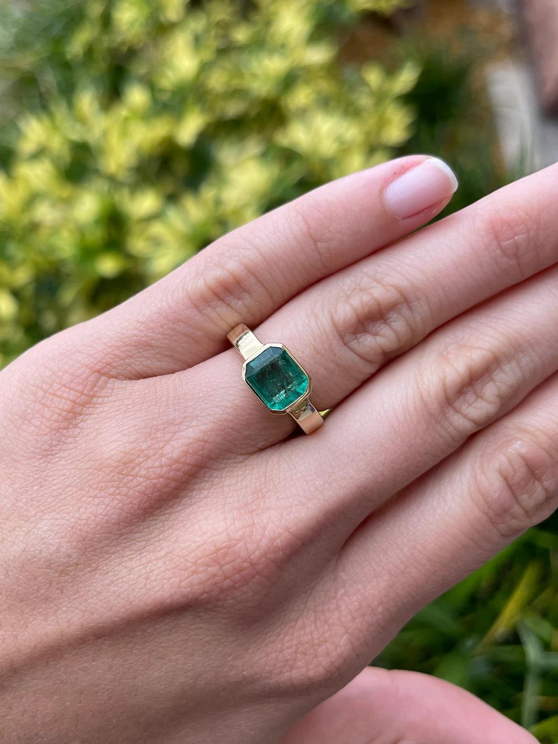 Displayed is a stunning East to West emerald solitaire engagement or right-hand ring in 14K yellow gold. This gorgeous solitaire ring carries a 2.40-carat emerald in a bezel setting. Fully faceted, this gemstone showcases excellent shine and