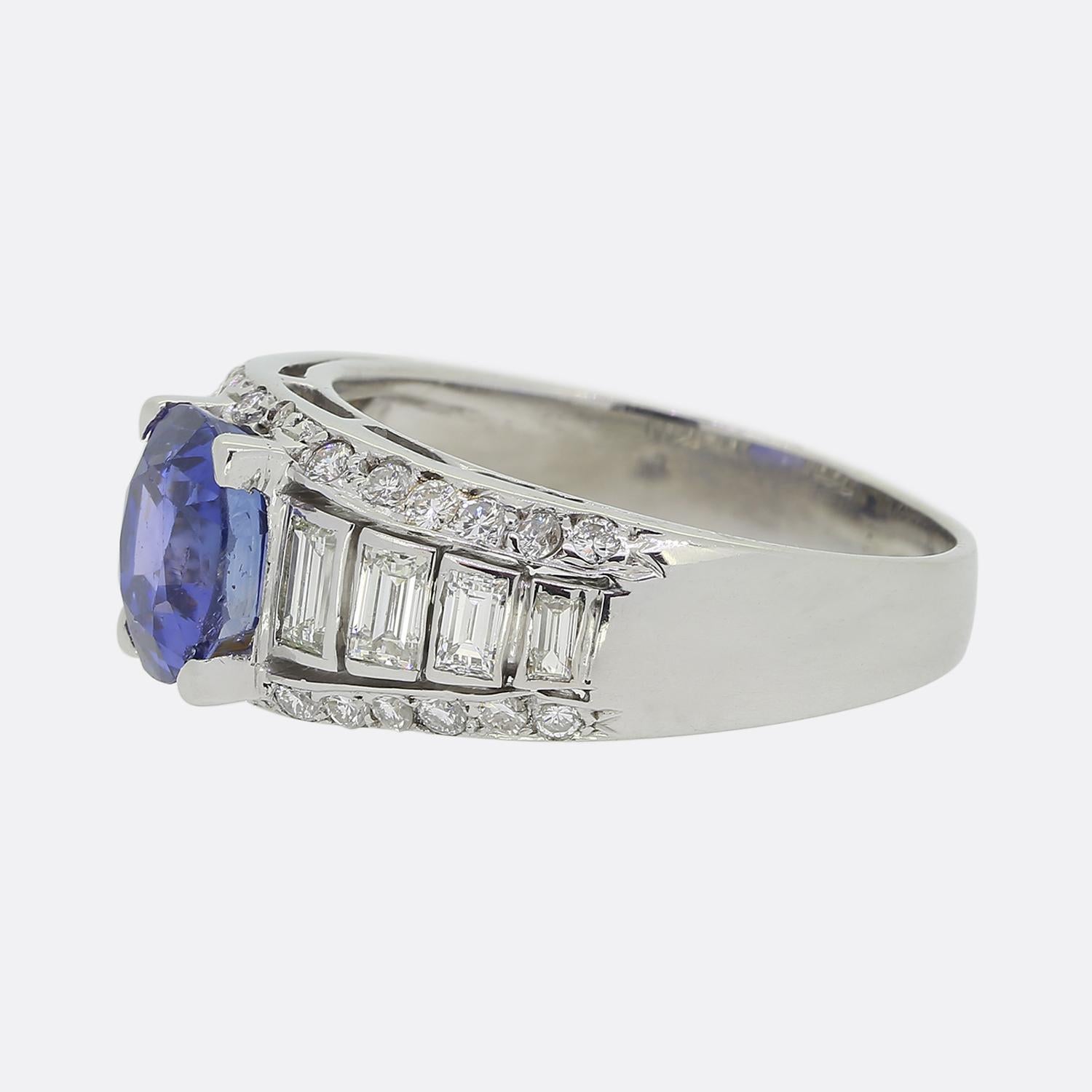 Here we have a delightful sapphire and diamond cluster ring. A single cushion cut sapphire of Ceylon origin boasting a wondrous cornflower blue colour tone sits slightly risen at the centre of the face. This principal stone is then flanked on either